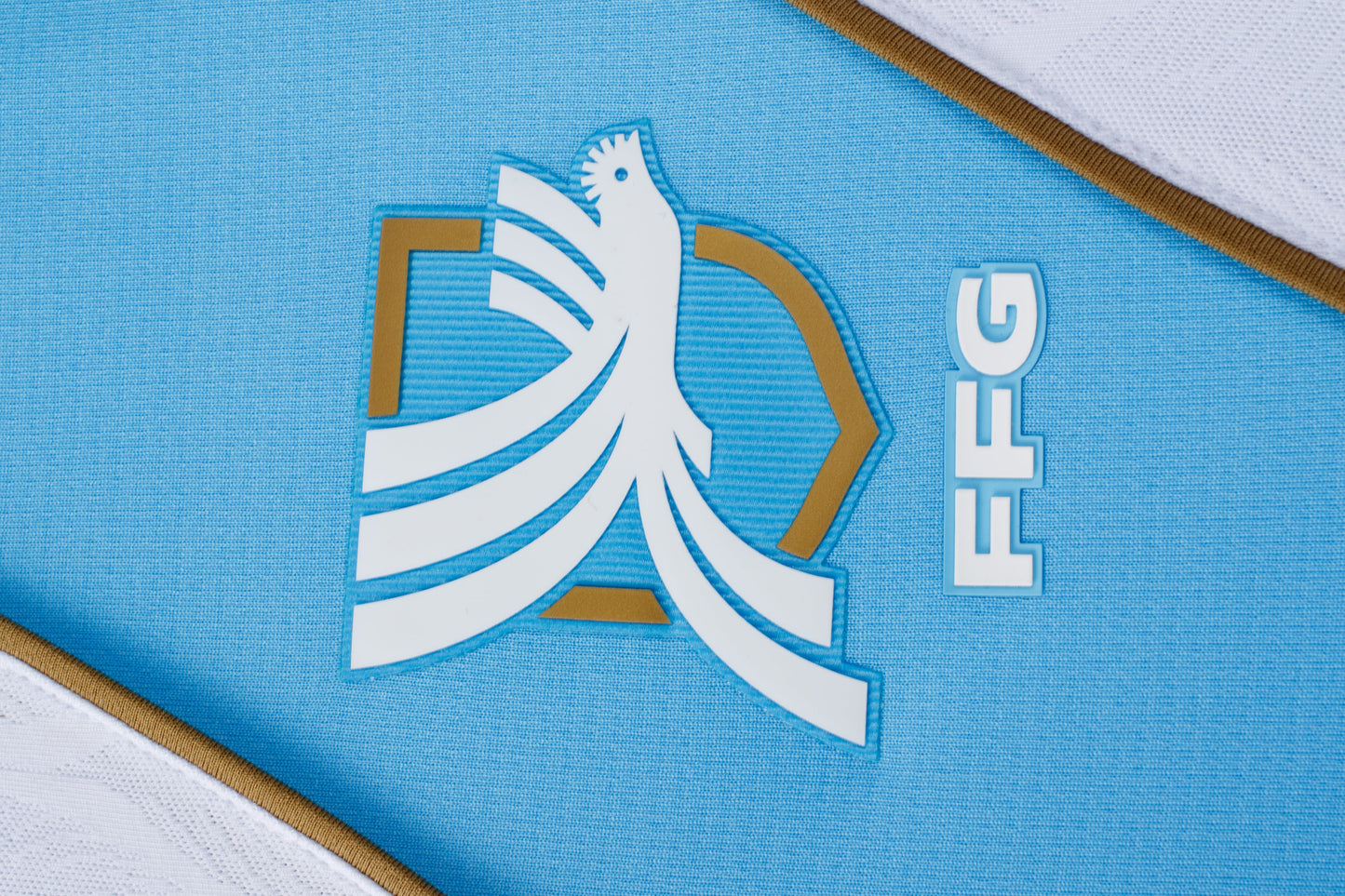 Close-up of a white rooster logo with gold accents and "FFG" text on light blue athletic fabric. White and gold trim is visible at the corners.