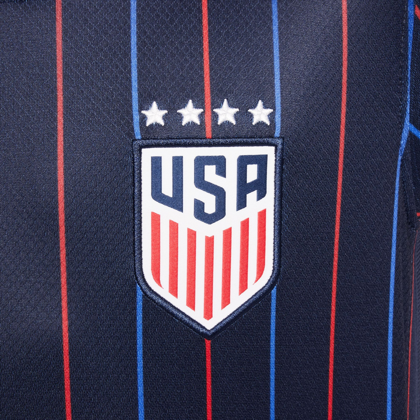 Close-up of a dark blue soccer jersey with red and blue vertical pinstripes, featuring the US national team shield crest and four white stars above it.