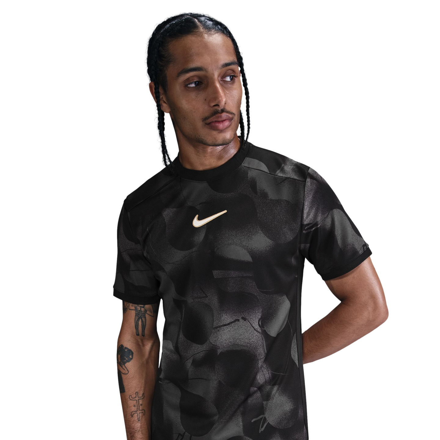 A man with braided hair wears a black patterned Nike t-shirt and looks to the side against a white background.