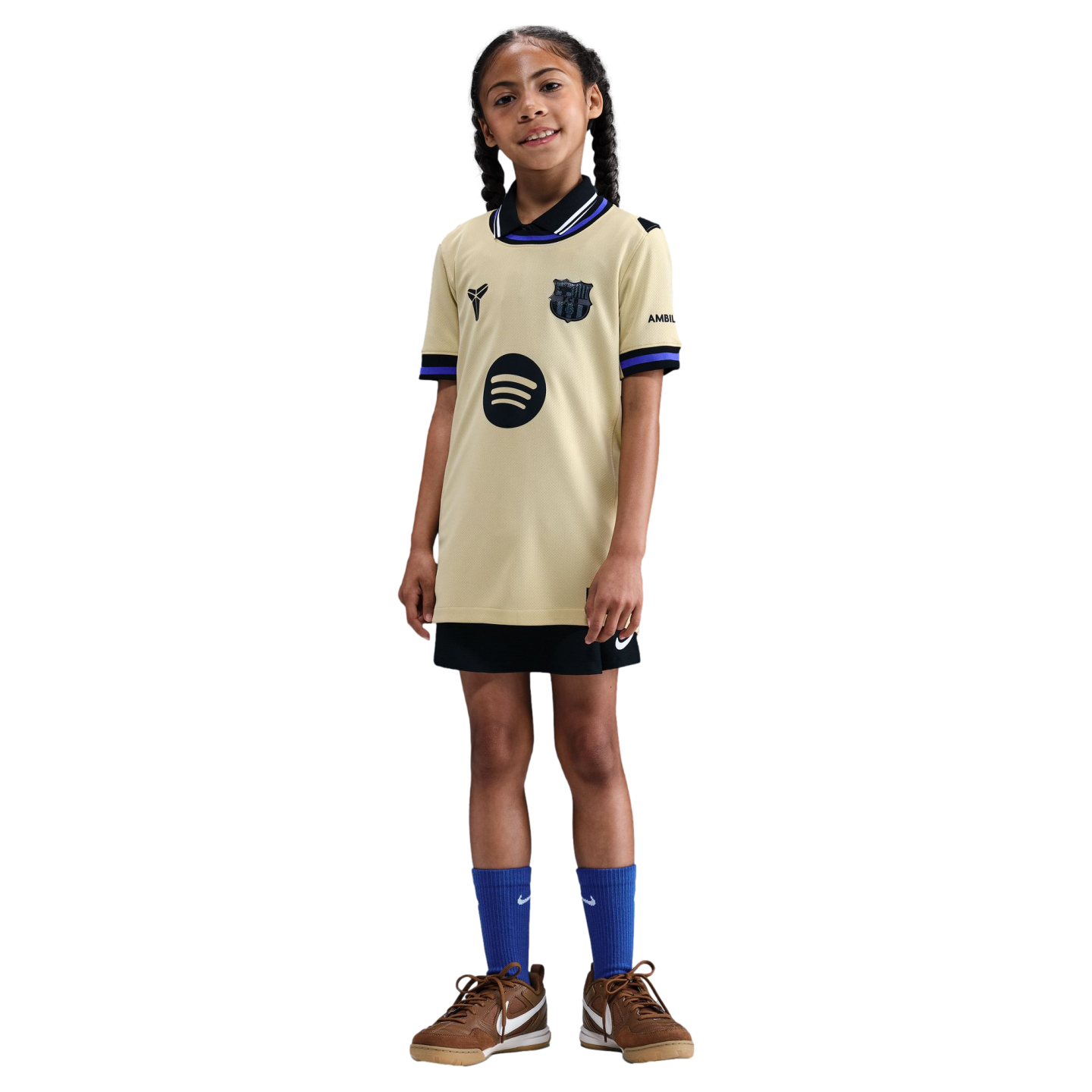 Young girl posing in a gold FC Barcelona Mamba jersey, black shorts, blue socks, and brown sneakers against a black background.