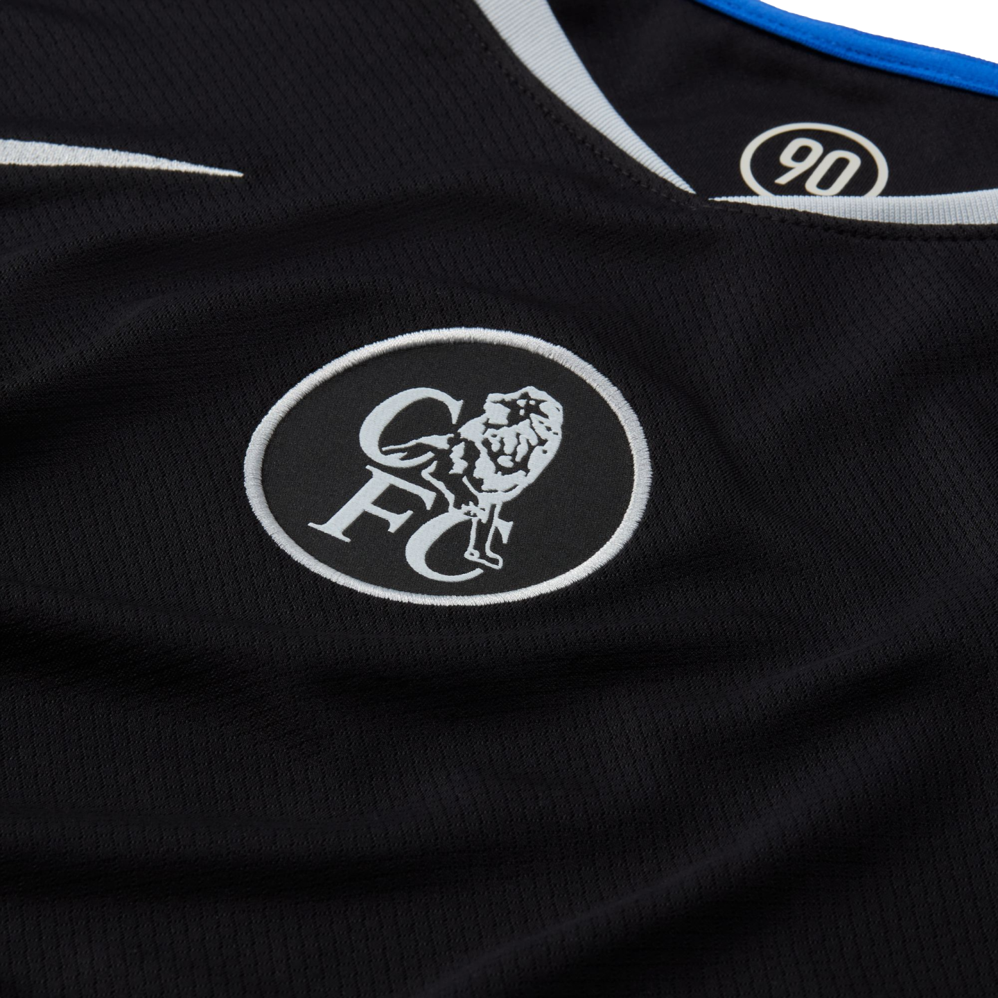 Close-up of a black Chelsea FC jersey with a white embroidered circular lion crest and "CFC" lettering. The collar features blue and white trim and a "90" logo inside.