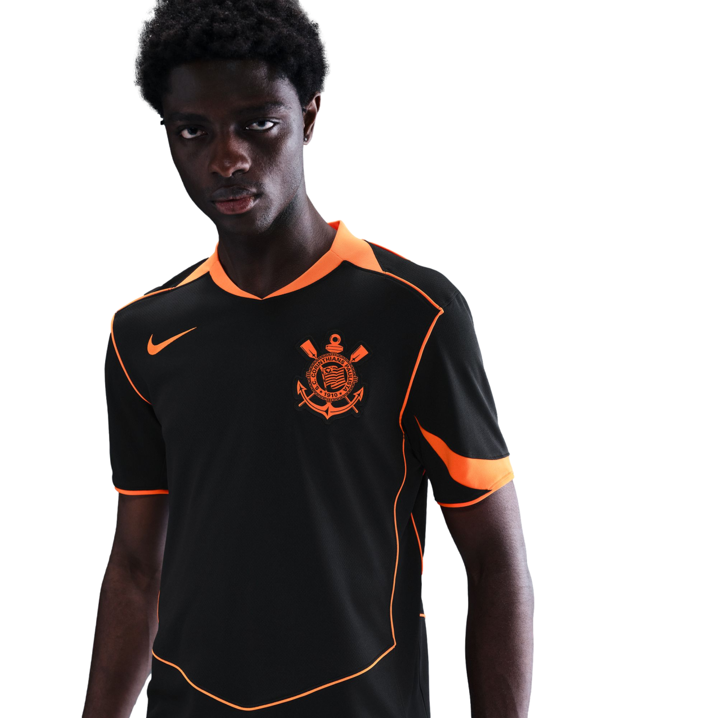 A Black man wearing a black soccer jersey with bright orange trim and a Corinthians crest against a black background.