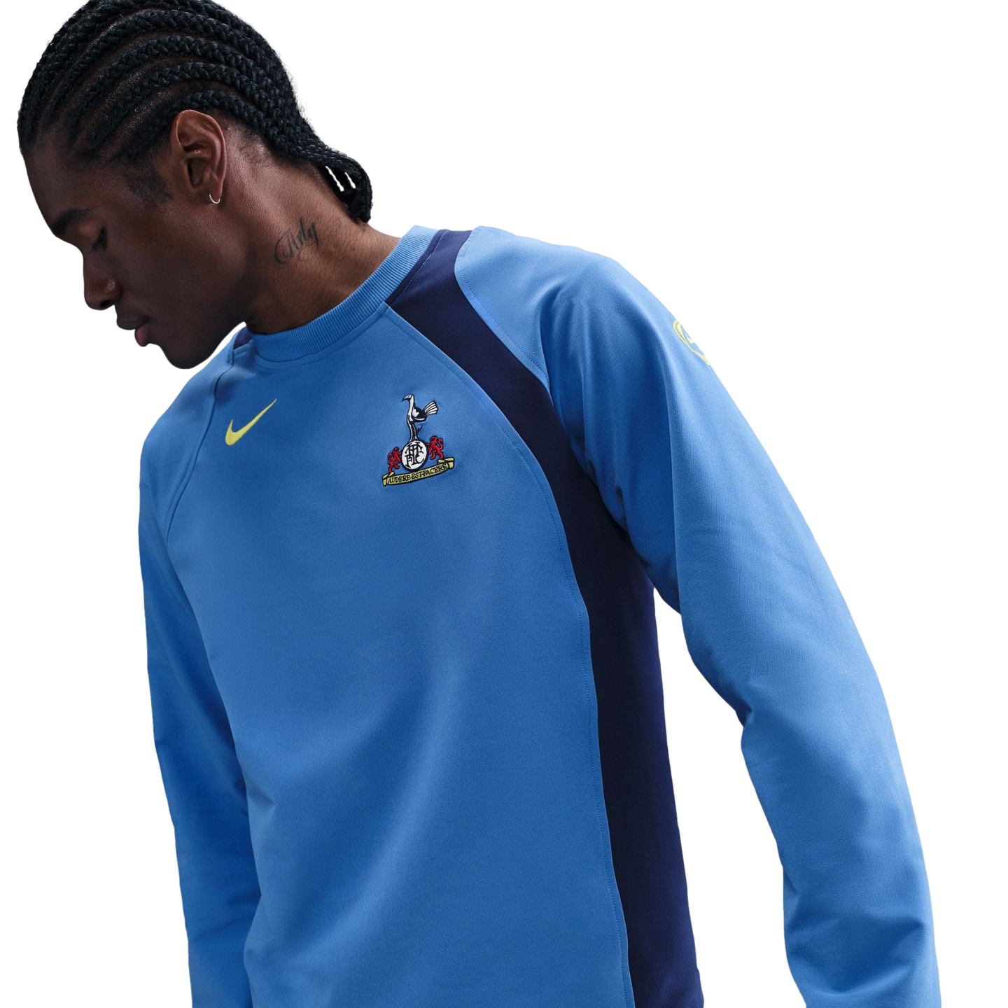 A man with braided hair and a neck tattoo wears a light blue Nike Tottenham Hotspur training shirt with navy blue side panels, looking down against a black background.
