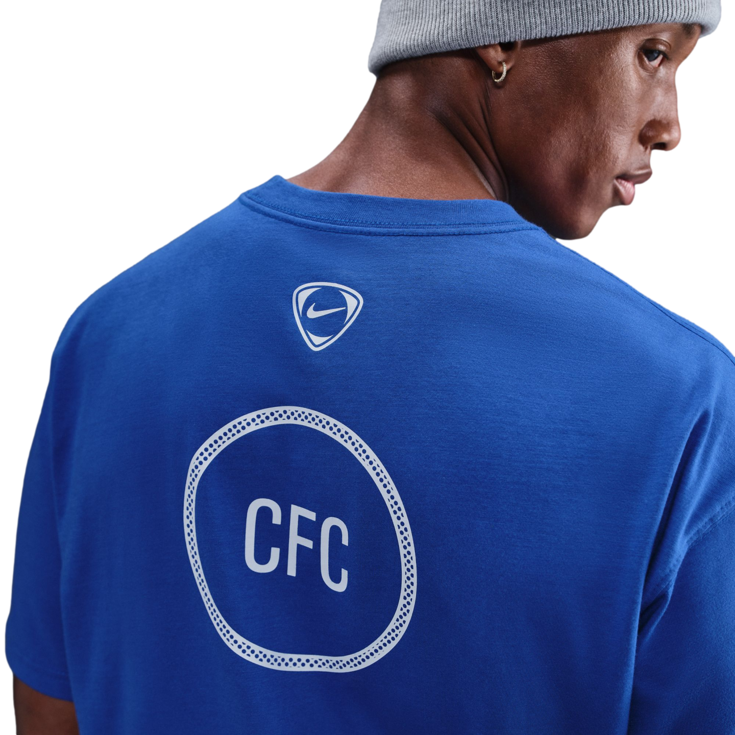 Back view of a man in a blue Chelsea FC t-shirt and grey beanie, showing a Nike logo and "CFC" inside a white patterned circle on the shirt.
