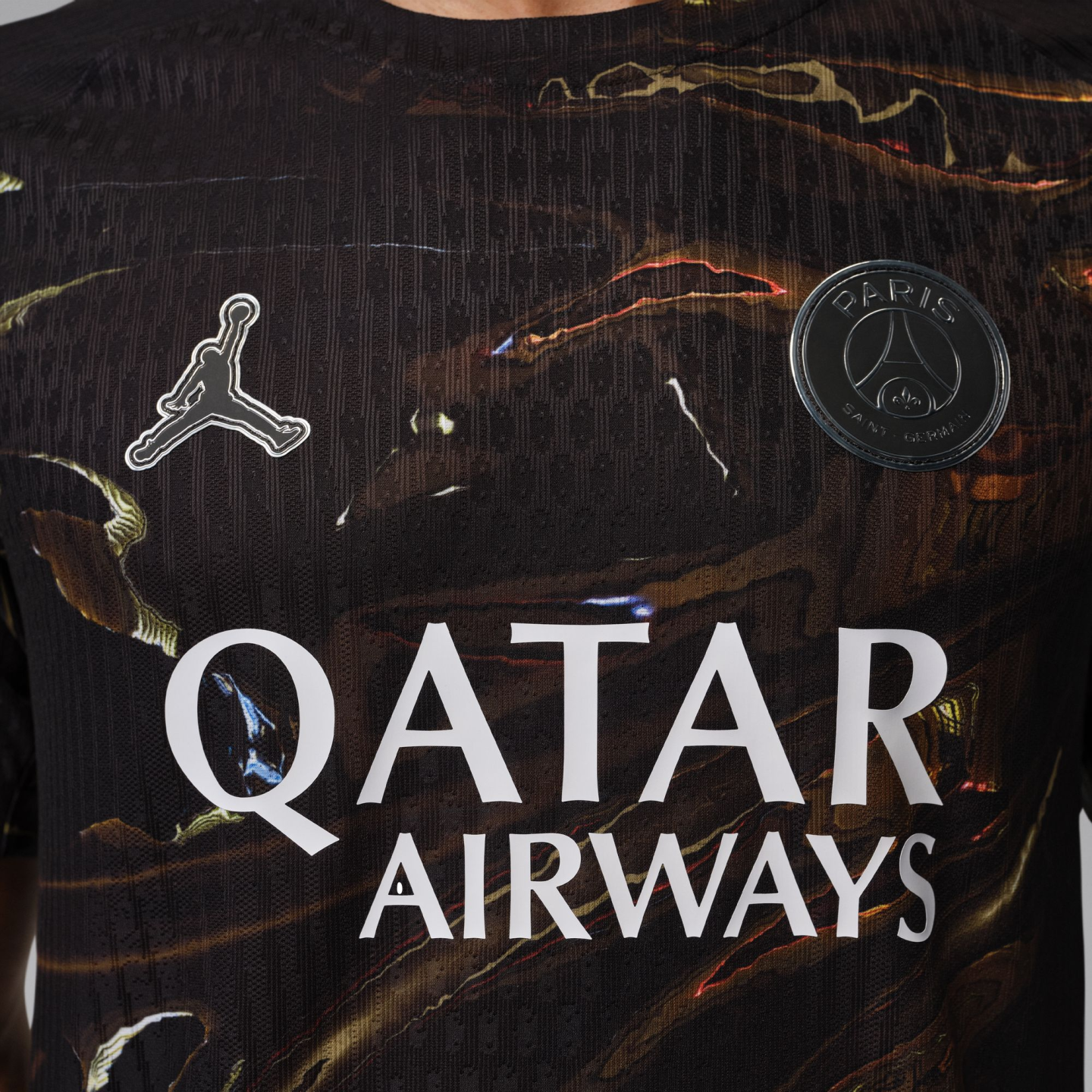 A close-up of a black Paris Saint-Germain soccer jersey with a brown and gold marble-like pattern. It features the white Jordan Brand "Jumpman" logo on the right chest, a black and silver PSG crest on the left chest, and the "QATAR AIRWAYS" sponsor logo i