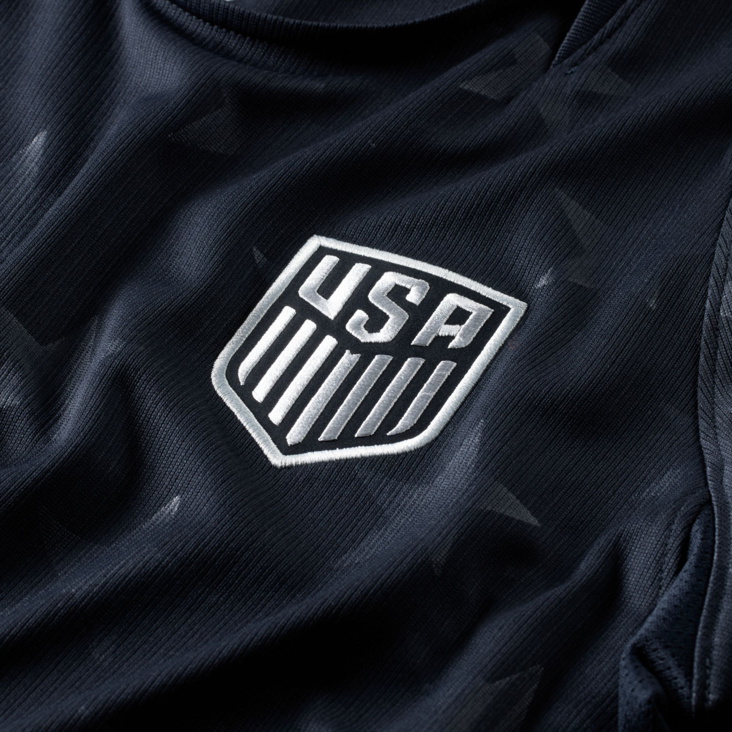 Close-up of a navy blue US soccer jersey with a metallic silver USA crest on textured fabric featuring subtle star patterns.
