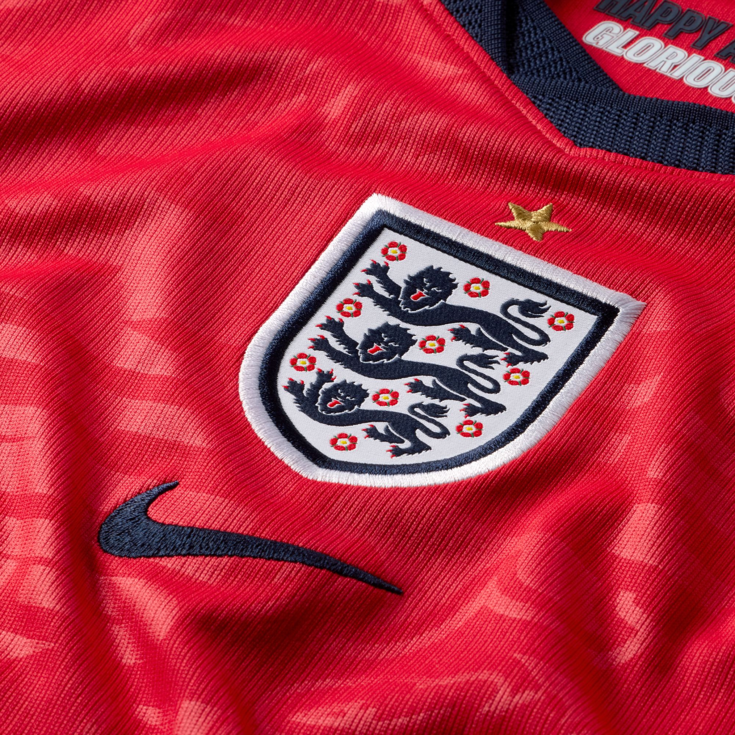 Close-up of a red England football jersey featuring the embroidered Three Lions crest, a dark blue Nike swoosh, and a single gold star above the badge on textured fabric.