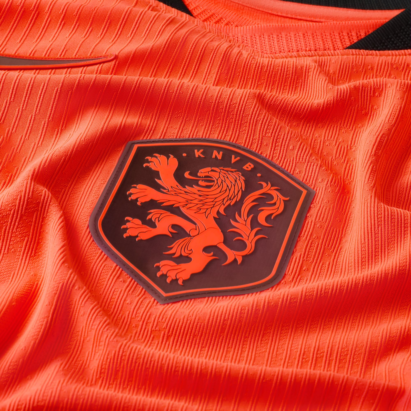 A close-up of a bright orange textured soccer jersey featuring the KNVB crest. The dark, shield-shaped patch displays an orange lion in a rampant pose with the letters "K N V B" above it. The jersey's fabric has a distinctive vertical ribbed pattern.