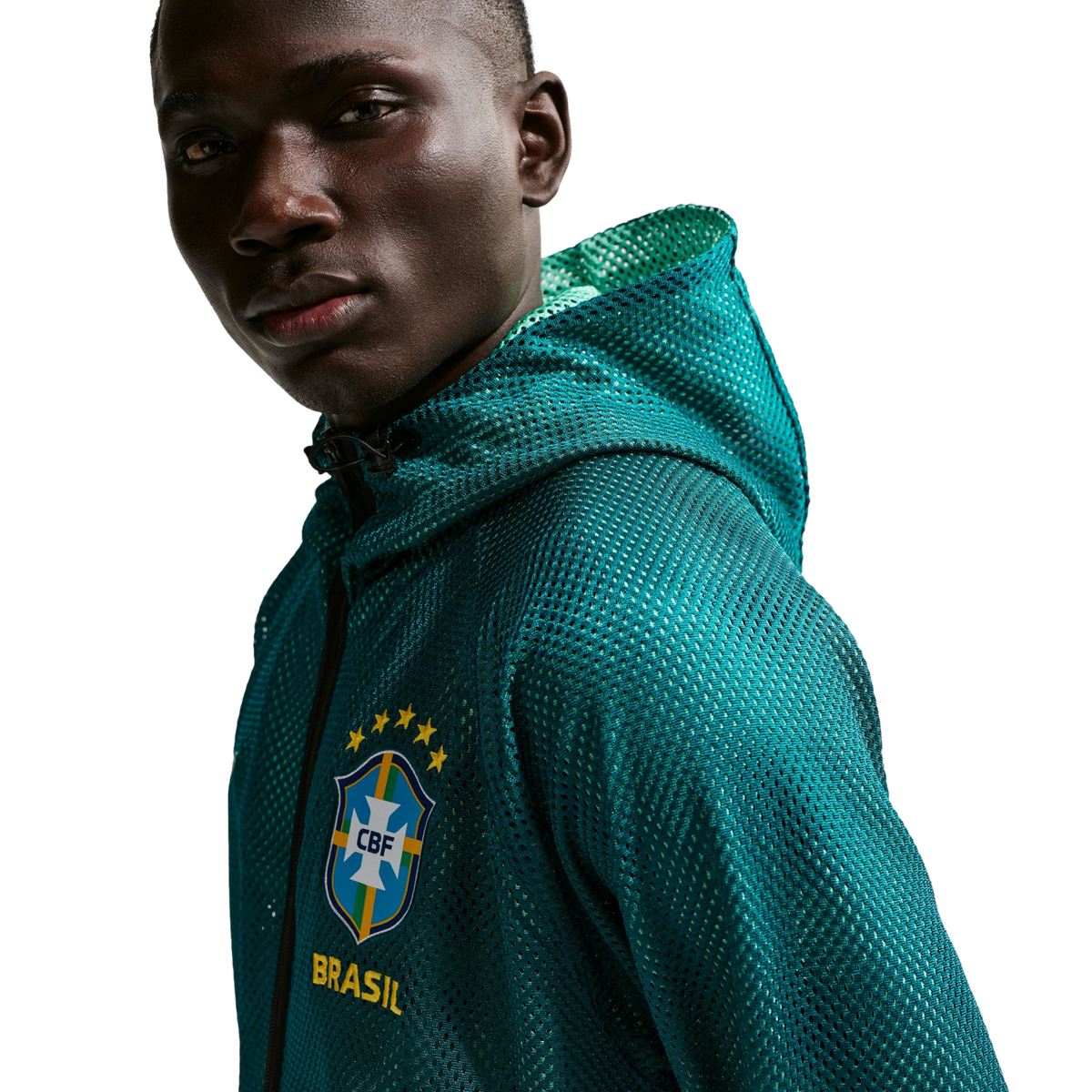A close-up shot of a man with dark skin looking towards the camera against a solid black background. He is wearing a teal, mesh, zip-up hoodie with the Brazilian football confederation (CBF) crest on the chest, featuring five yellow stars above it and the