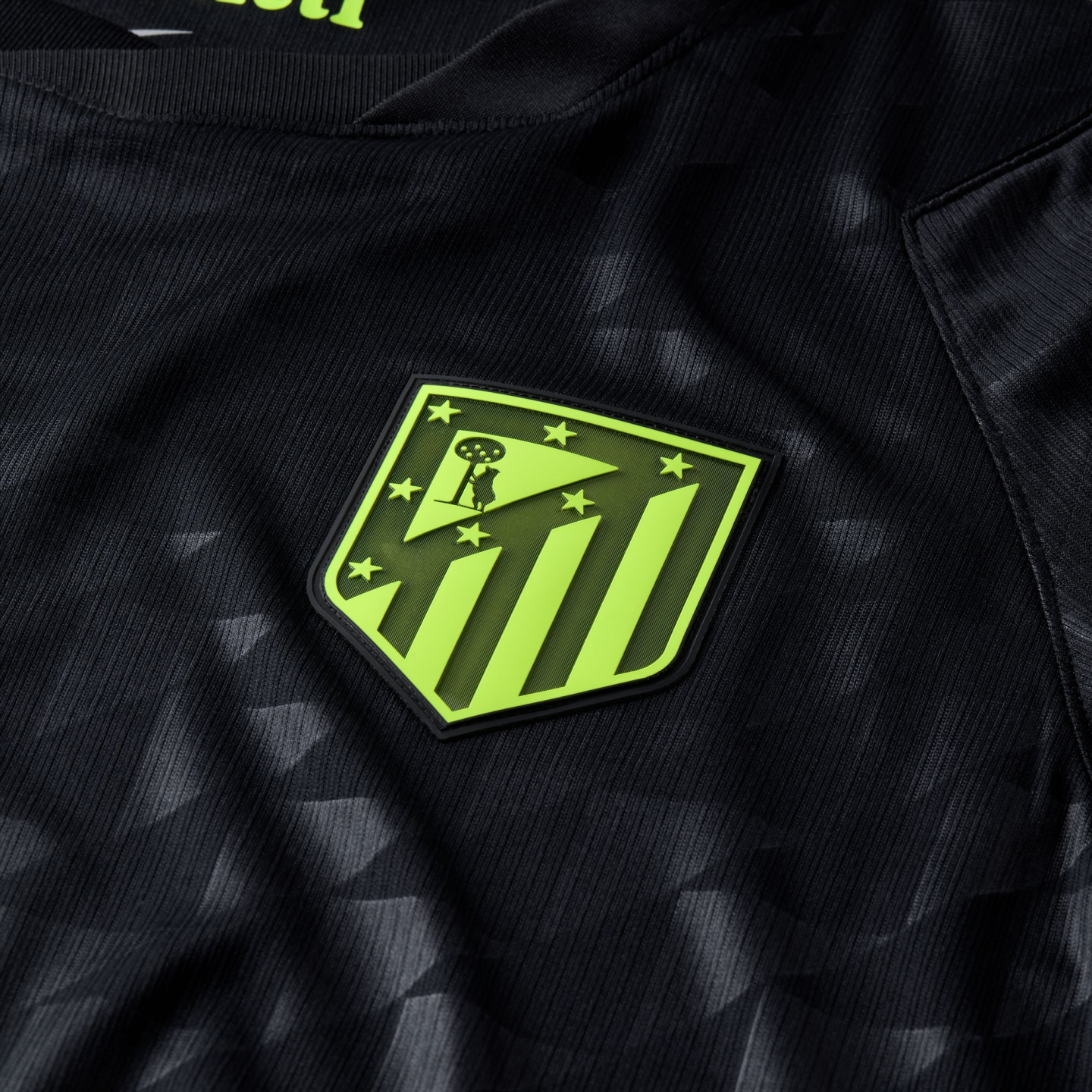 Close-up of a black soccer jersey with a lime green Atlético Madrid crest. The fabric features a subtle tonal geometric pattern.