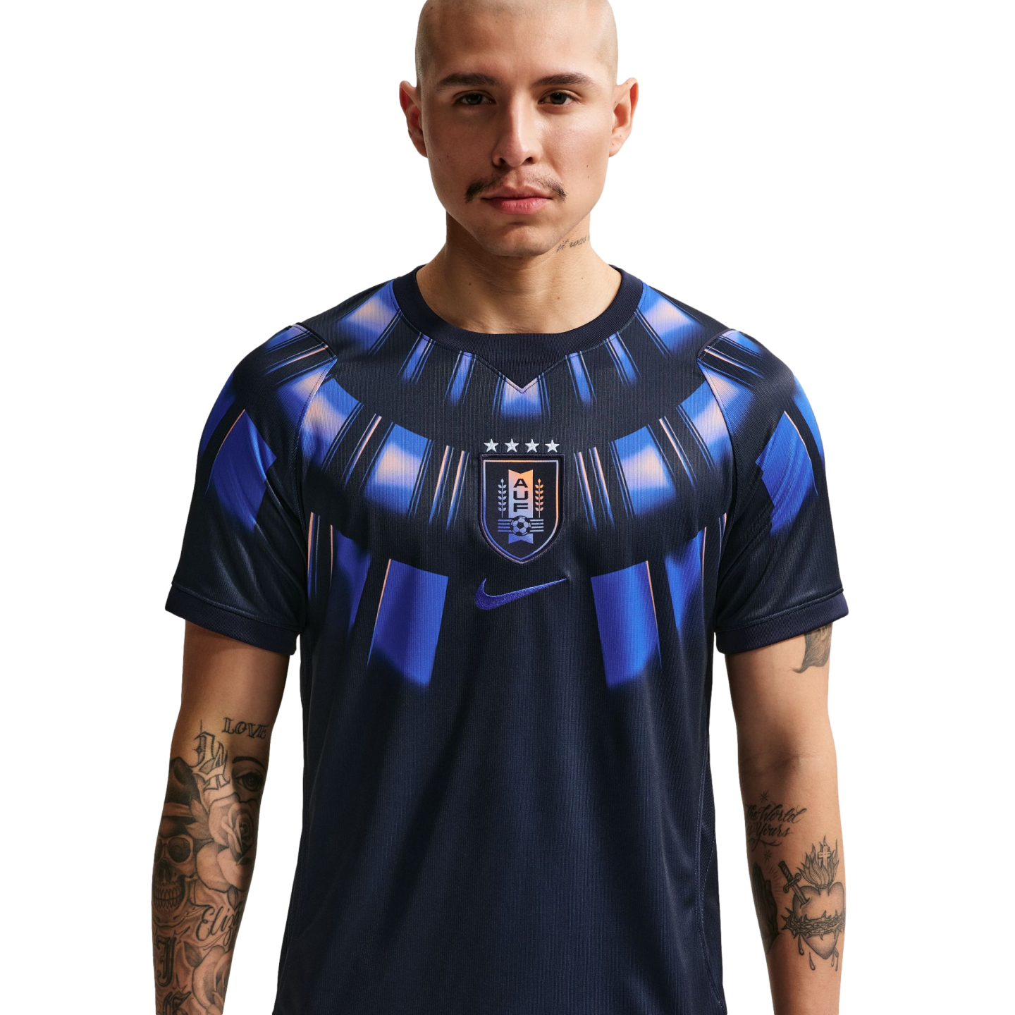 A waist-up studio portrait of a bald man with arm tattoos wearing a dark navy Uruguay national team soccer jersey featuring a blue and orange sunburst pattern around the collar and the AUF crest in the center against a solid black background.