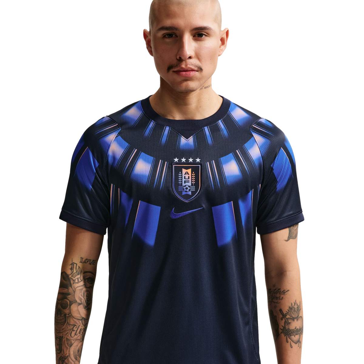 A waist-up studio portrait of a bald man with arm tattoos wearing a dark navy Uruguay national team soccer jersey featuring a blue and orange sunburst pattern around the collar and the AUF crest in the center against a solid black background.