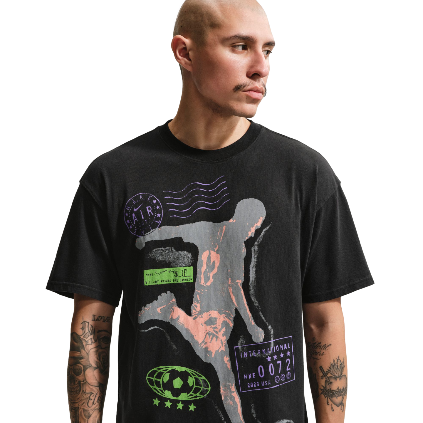 A man with a shaved head and mustache wears a black graphic t-shirt against a solid black background. The shirt features a large grey and peach-colored silhouette of an athlete in motion, surrounded by various purple and green sports-themed logos and text
