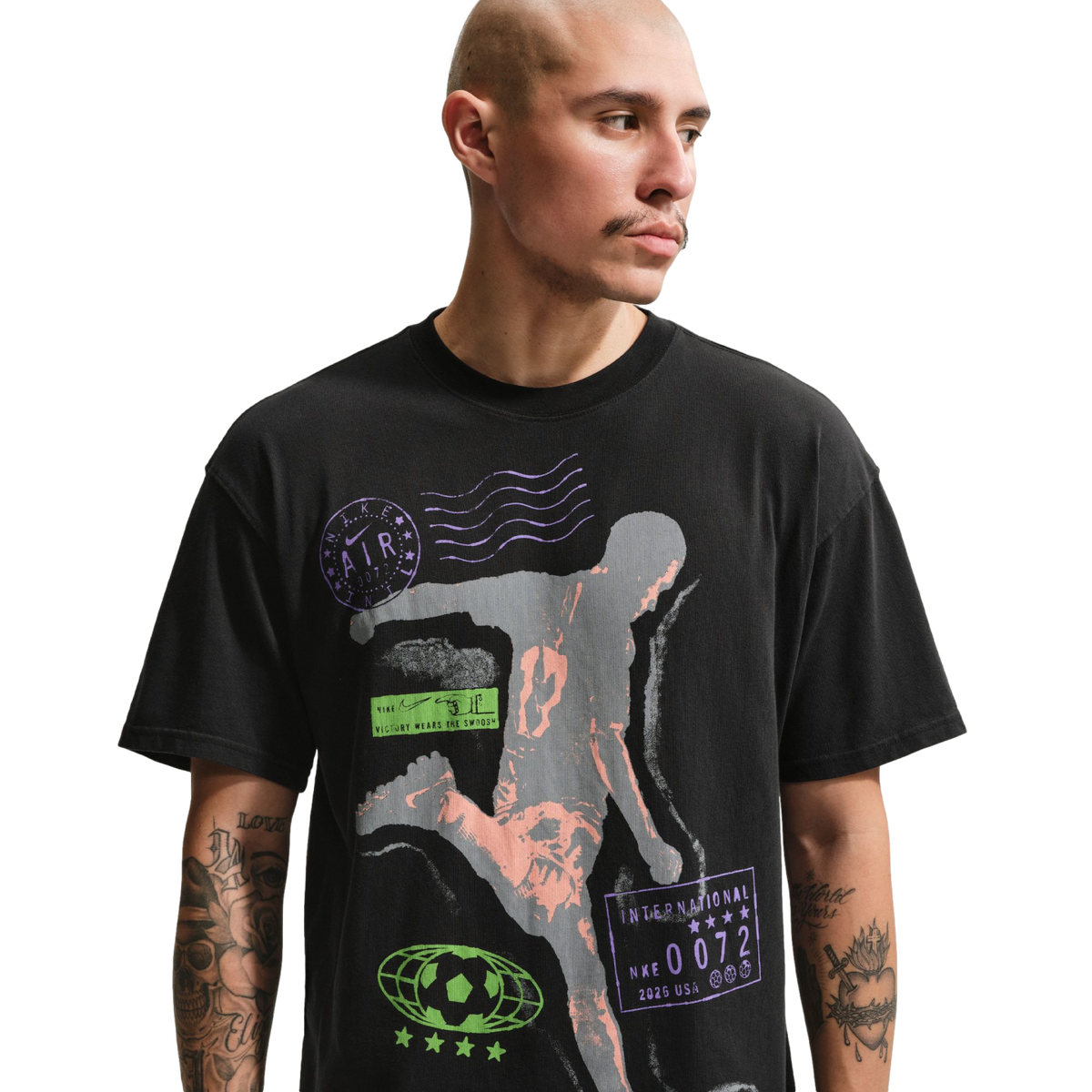 A man with a shaved head and mustache wears a black graphic t-shirt against a solid black background. The shirt features a large grey and peach-colored silhouette of an athlete in motion, surrounded by various purple and green sports-themed logos and text