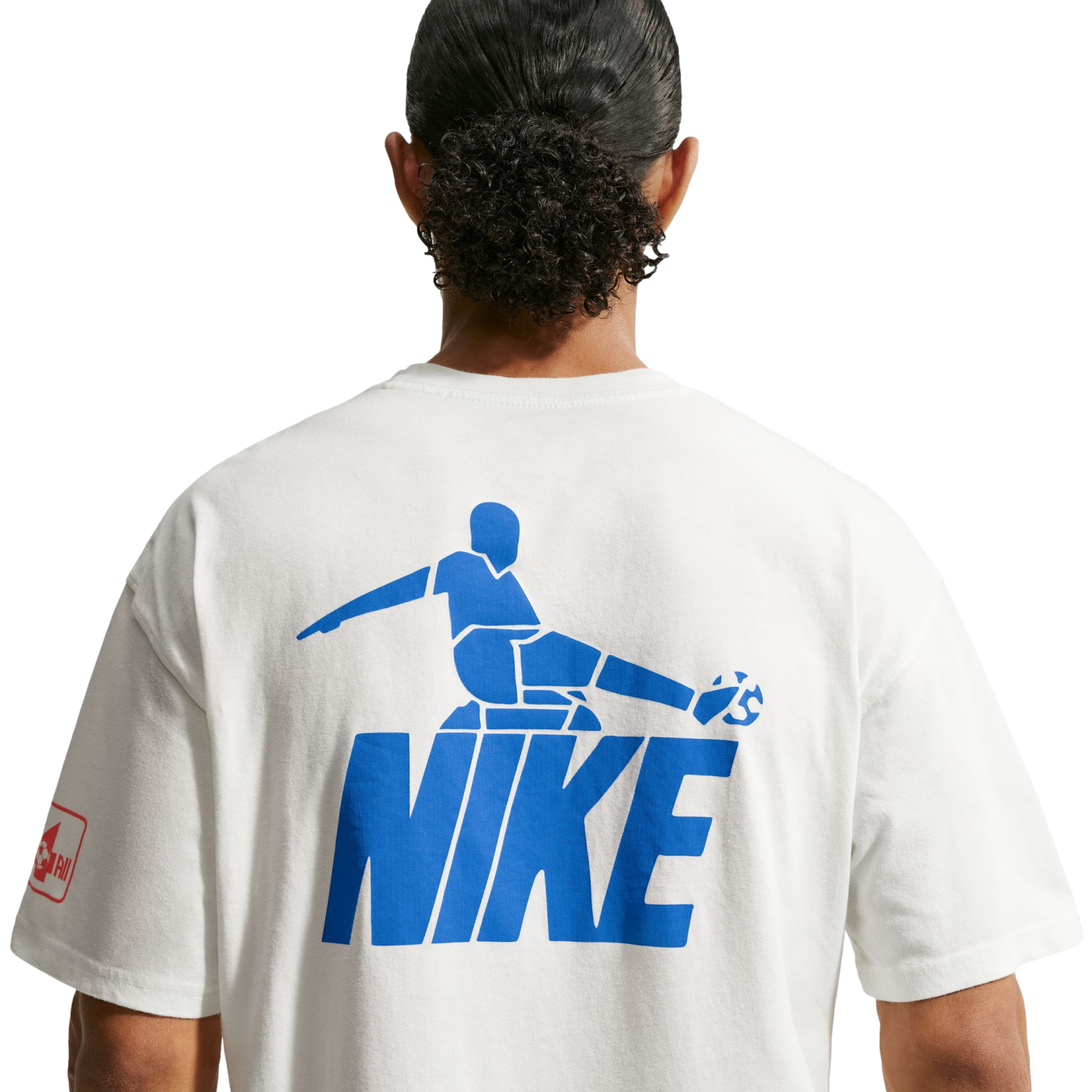 A back view of a person with dark, curly hair tied in a bun, wearing a white t-shirt against a black background. The shirt features a large blue graphic on the back consisting of a stylized soccer player kicking a ball, positioned above the word "NIKE" in