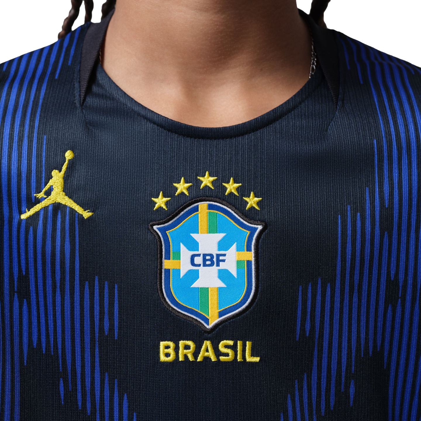 Close-up of a navy blue Brazil soccer jersey featuring a yellow Jumpman logo and the CBF crest with five stars above it, set against a background of textured blue vertical lines.