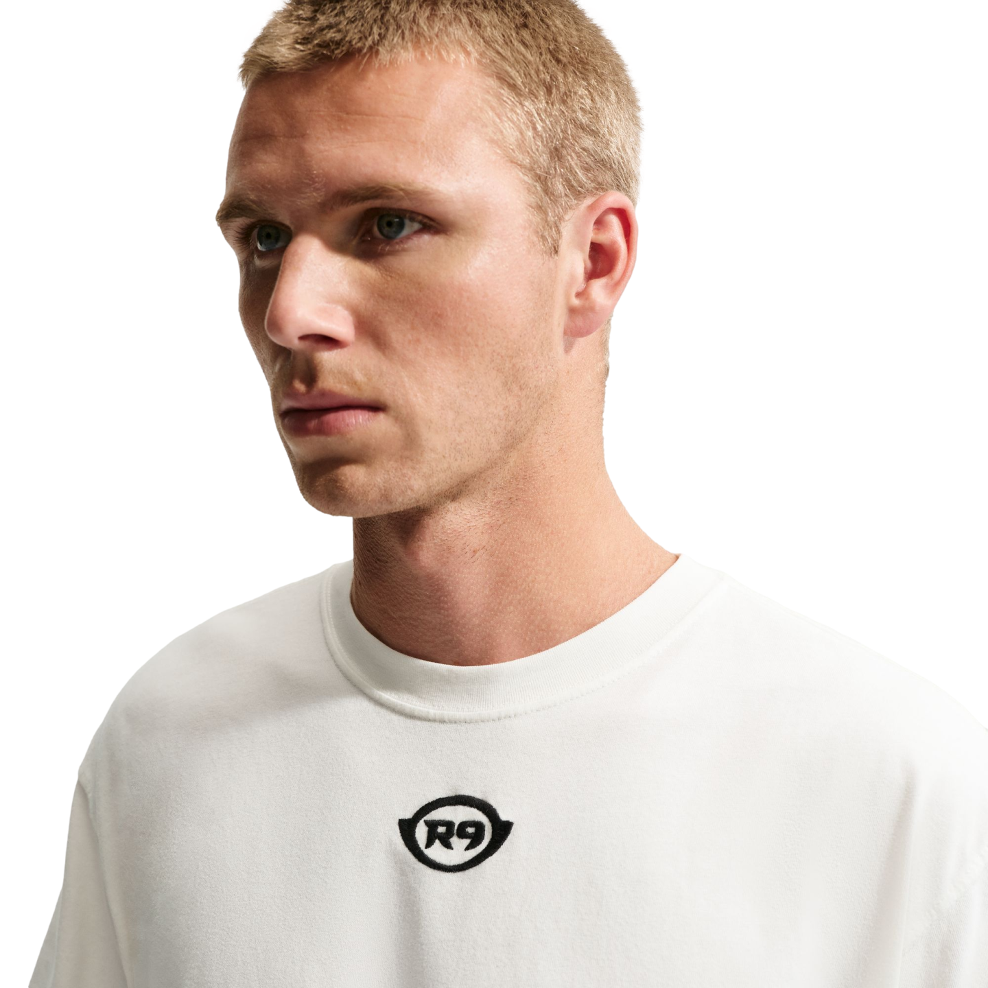 A medium shot of a man with short blonde hair wearing a white t-shirt with a small black "R9" logo in the center, set against a solid black background.