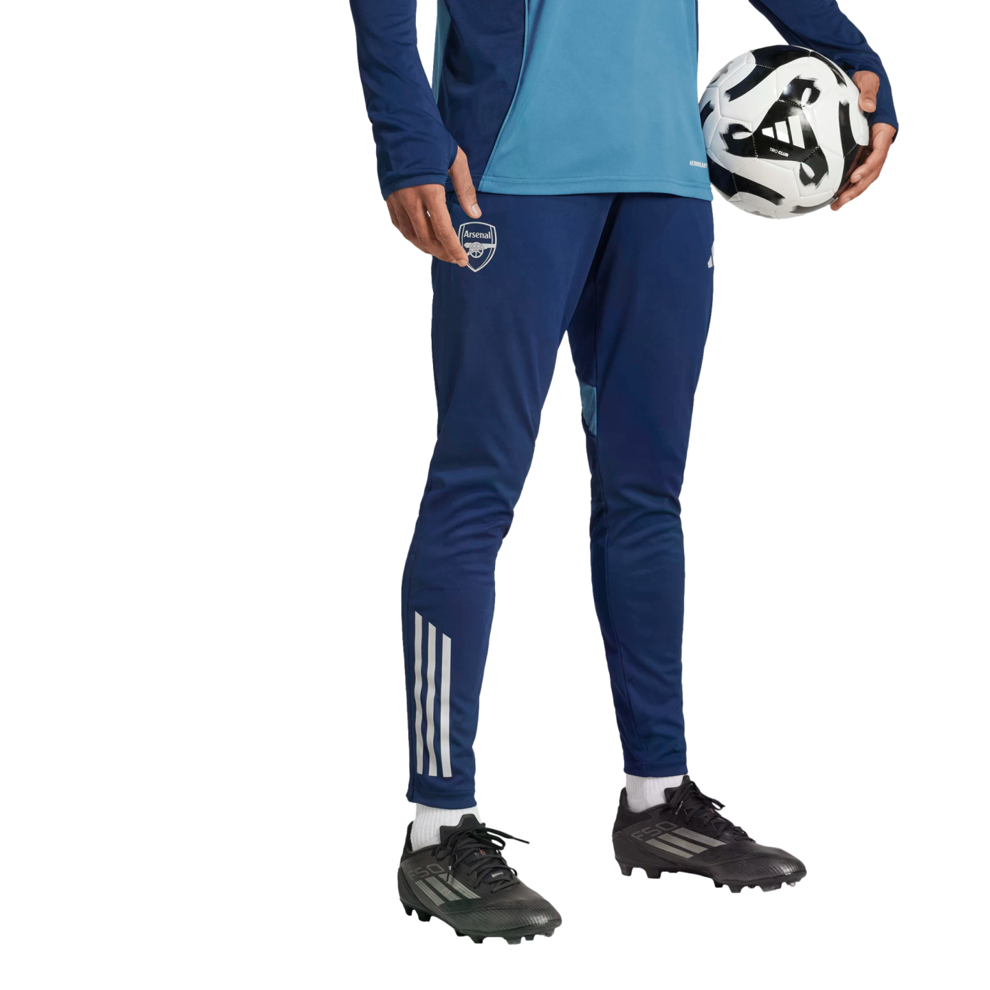 Person in blue Arsenal FC training gear and black soccer cleats holding a soccer ball against a black background.