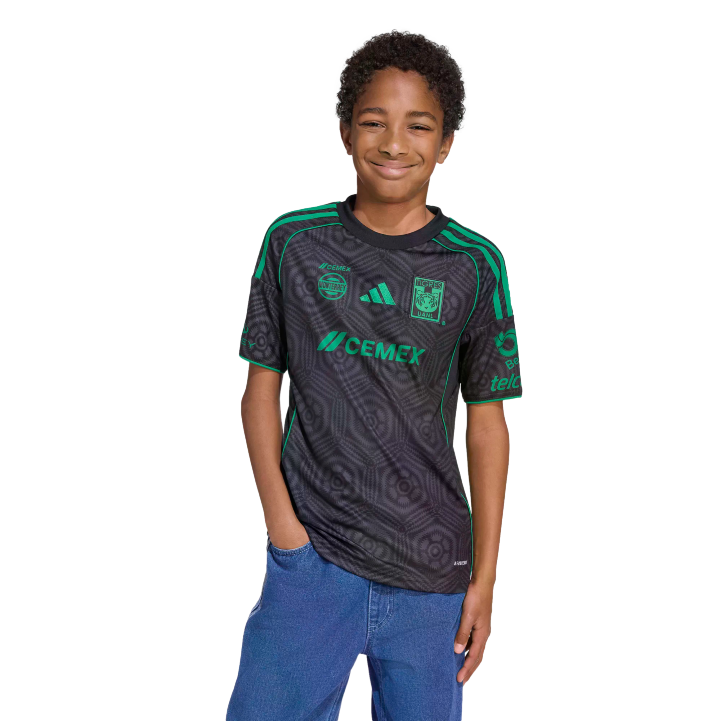 A smiling boy wearing a black and green Tigres UANL soccer jersey and blue jeans stands against a black background.