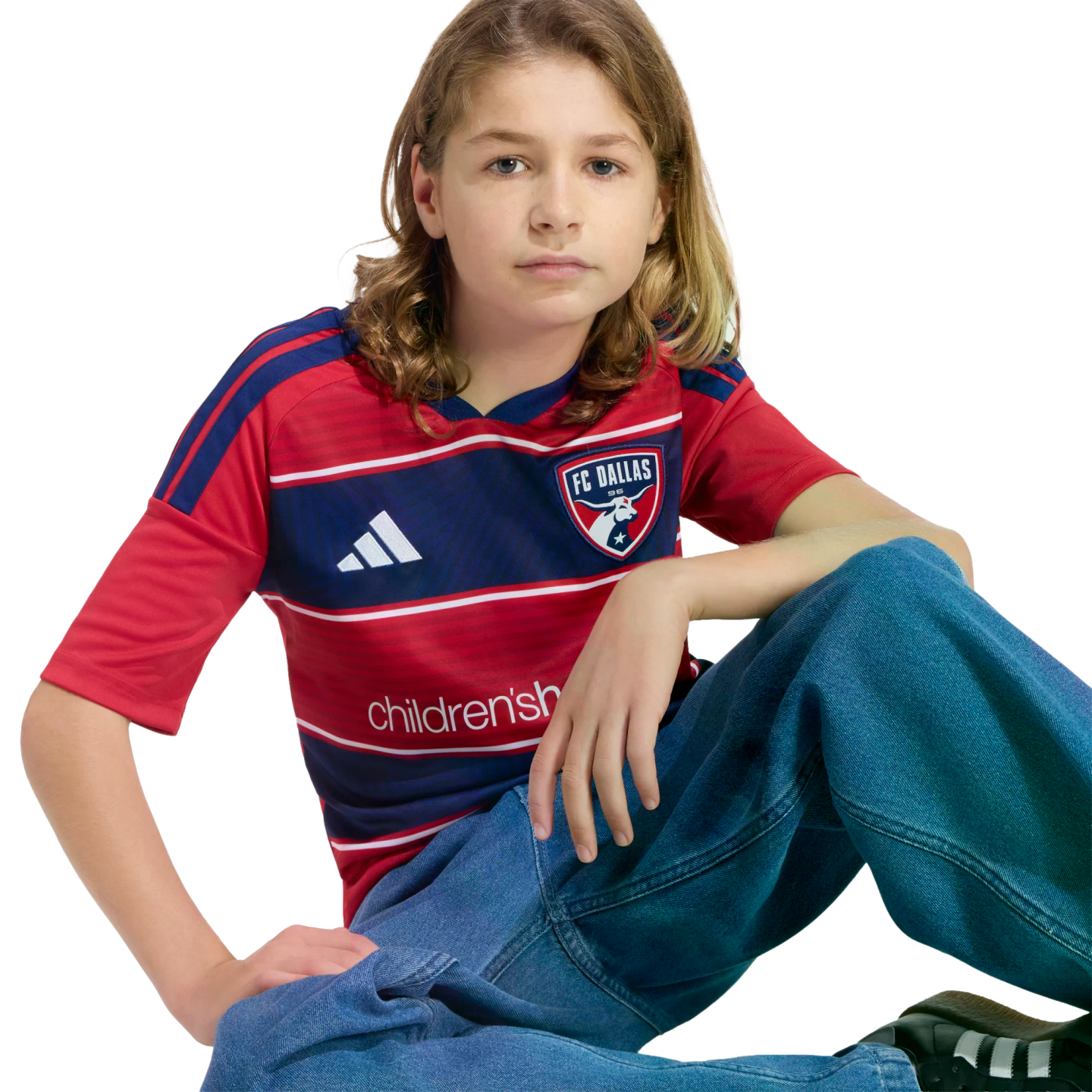 A young person with long hair sits in an FC Dallas jersey and baggy blue jeans against a black background.