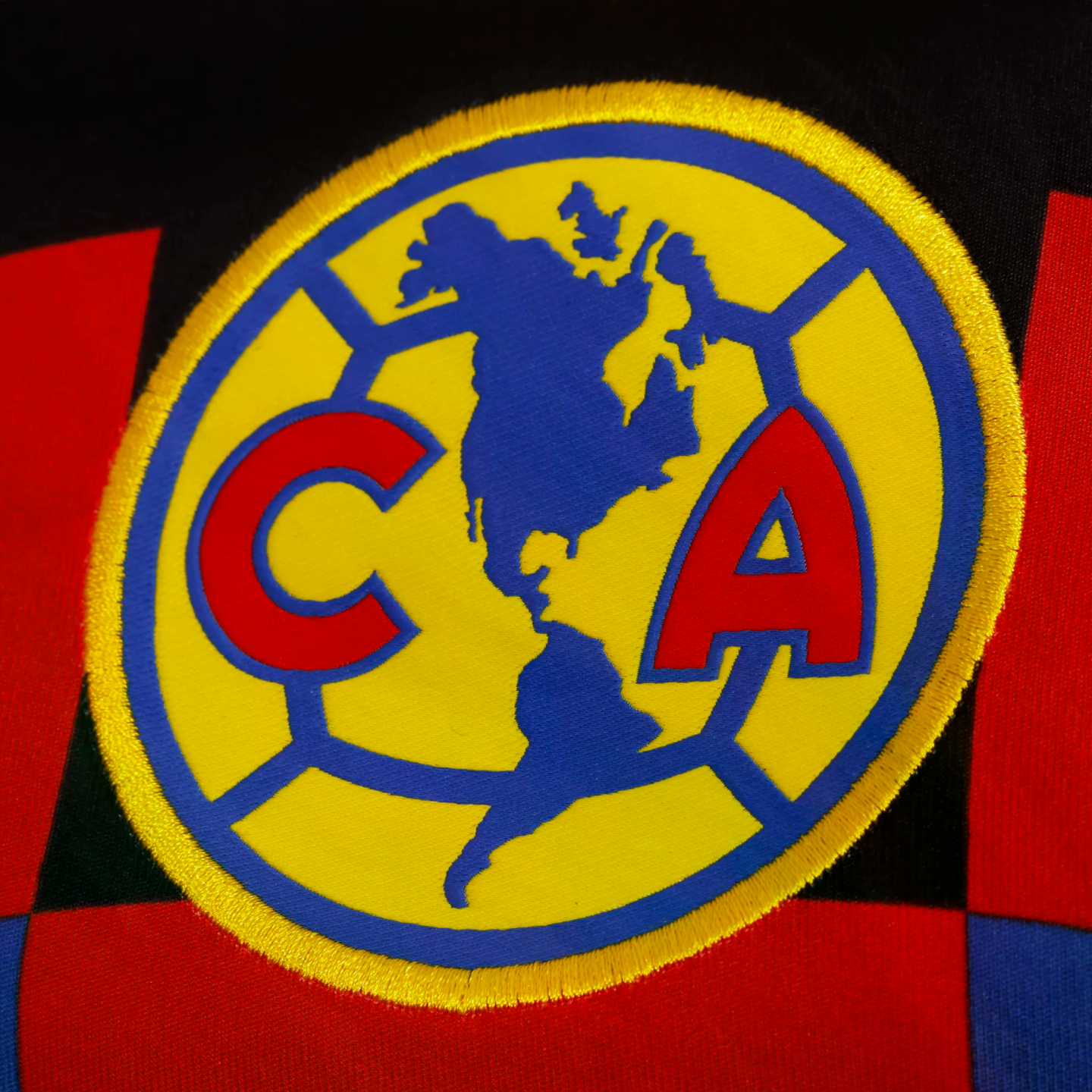 Embroidered Club América logo: a yellow circle with a blue map of the Americas and red 'C' and 'A' initials, set against a red and blue checkered fabric background.