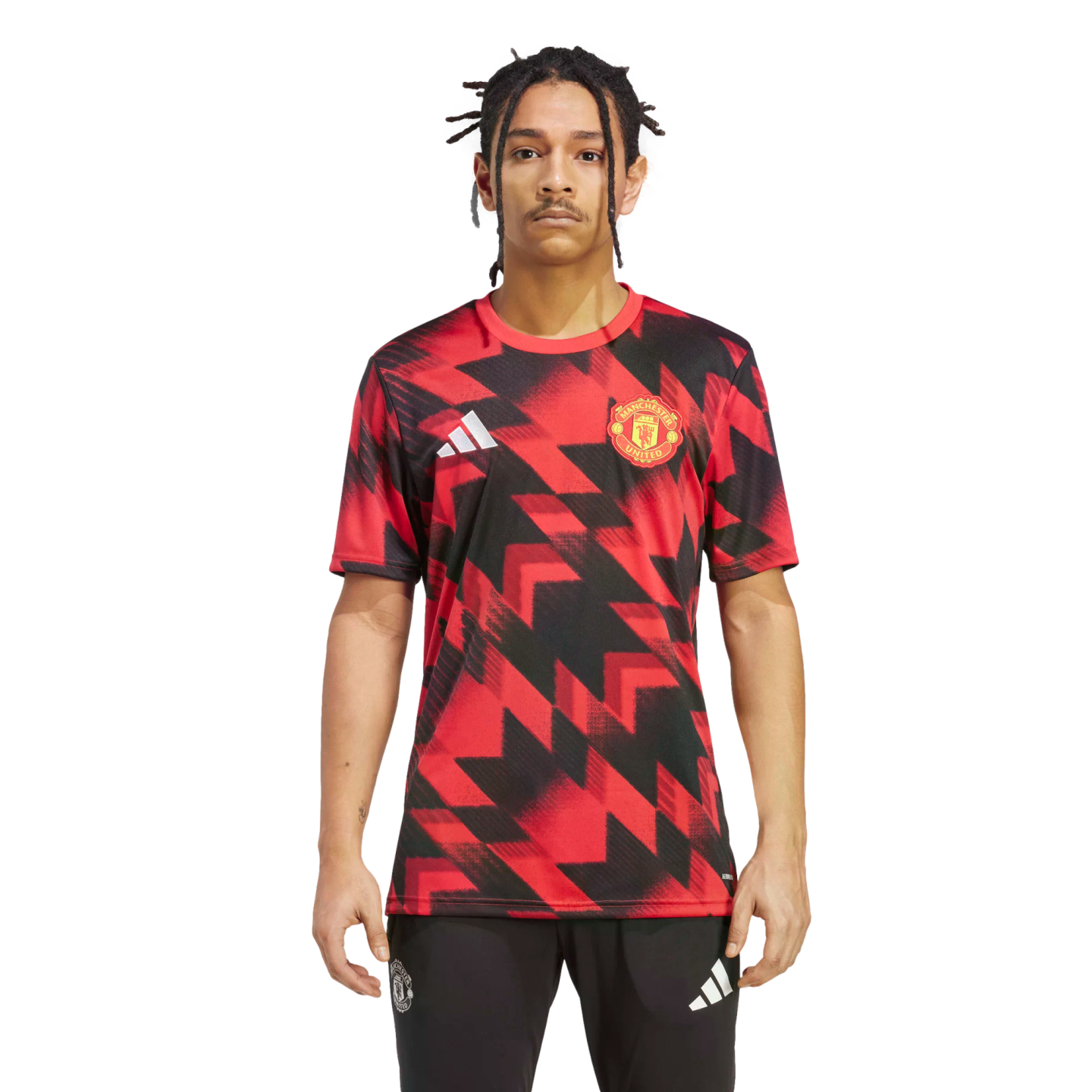 A man models a Manchester United training jersey featuring a red and black geometric pattern, paired with black Adidas joggers, against a plain white background.