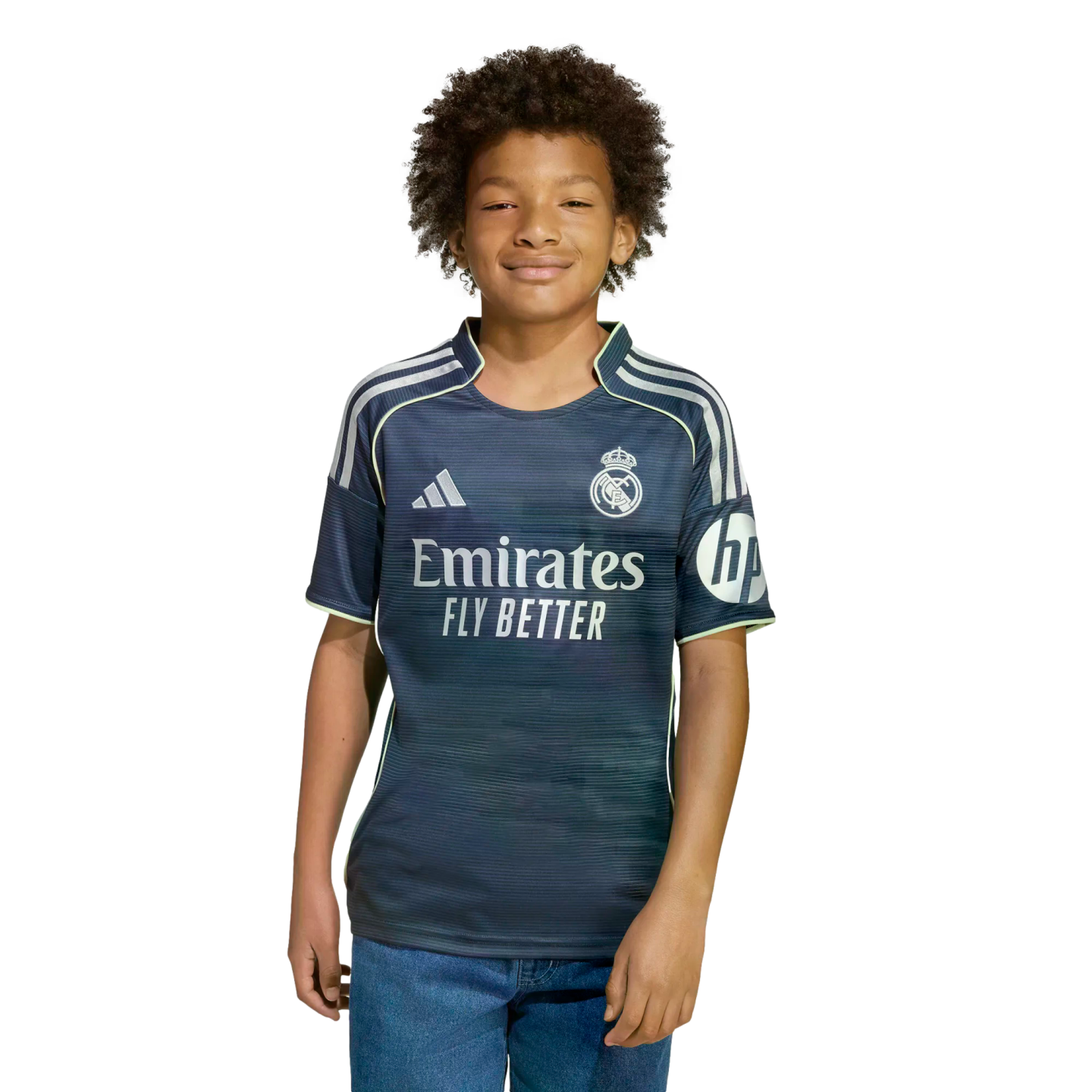 A boy with dark curly hair wearing a navy blue Real Madrid soccer jersey and blue jeans, smiling against a plain white background.