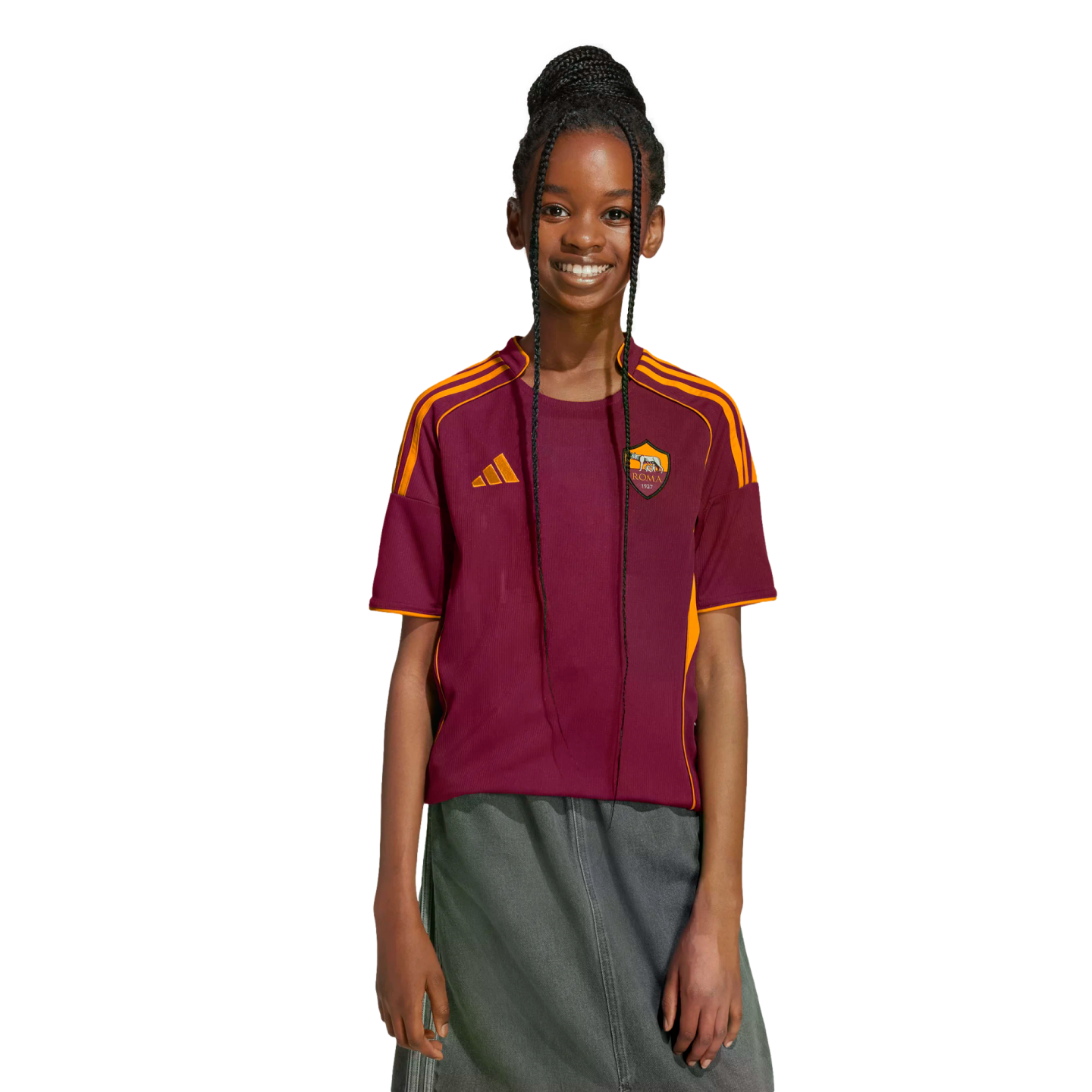 A young person with long braids smiles, wearing a maroon and orange AS Roma jersey and a grey skirt against a black background.