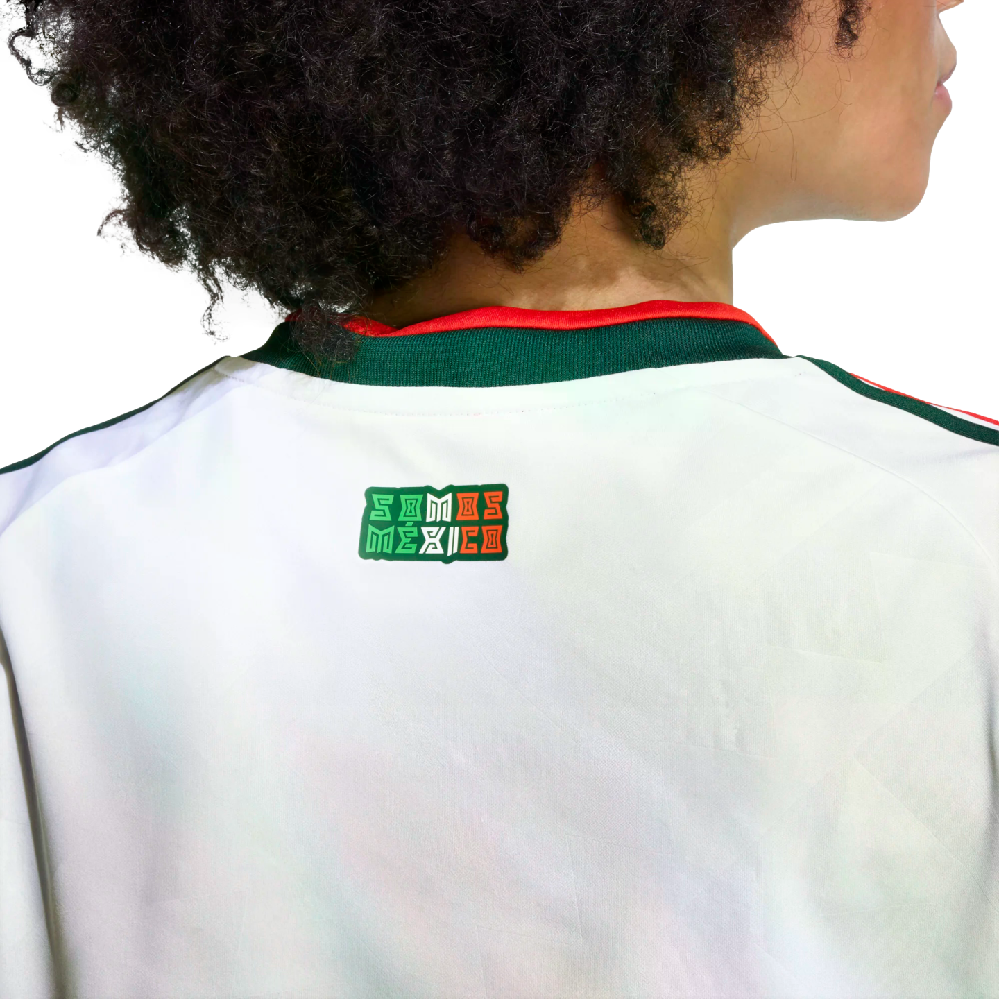 A person with dark curly hair from behind, wearing a white Mexico national team jersey. On the upper back, just below the green and red collar, is a small rectangular logo that reads "SOMOS MÉXICO" in the colors of the Mexican flag.