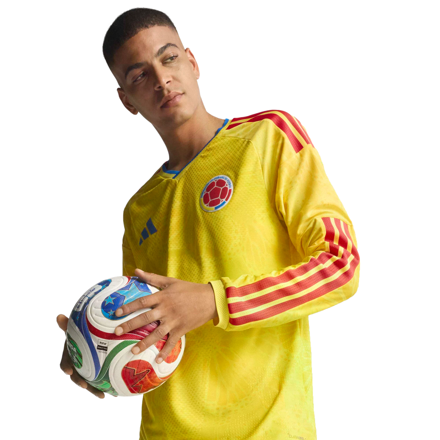 A man in a long-sleeved yellow Colombia soccer jersey with red stripes holds a colorful soccer ball against a solid black background.