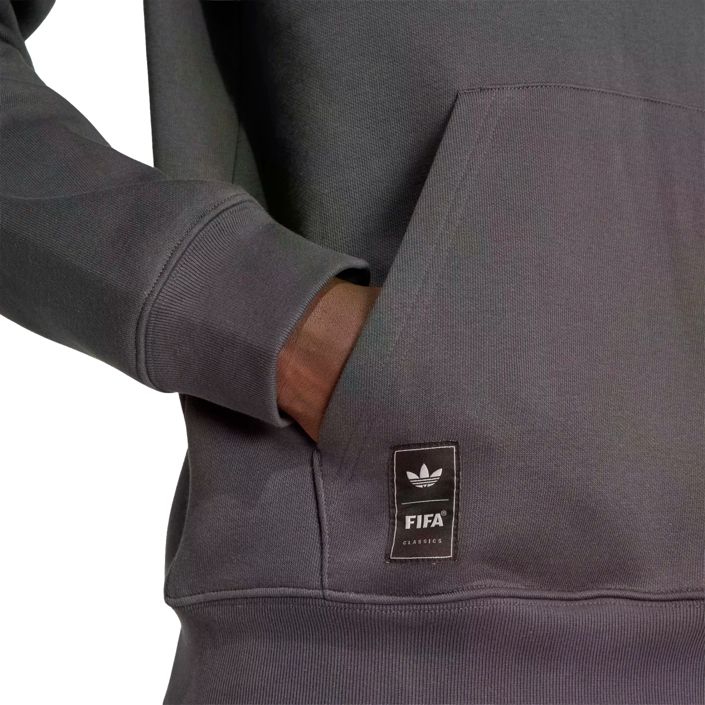 Close-up of a person's hand in a grey hoodie pocket. A black patch on the pocket features the Adidas trefoil logo and "FIFA CLASSICS" text.