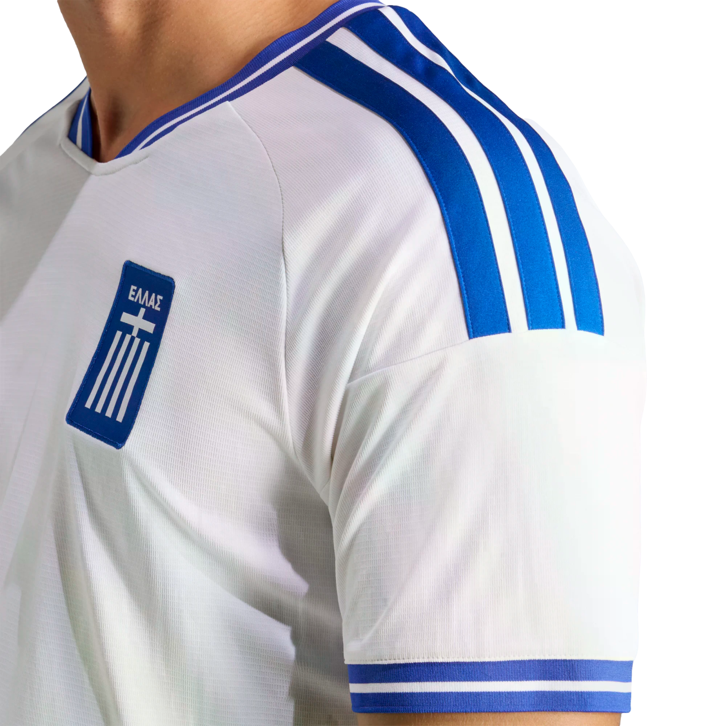 Close-up of a white athletic jersey with blue stripes on the shoulder and sleeve. A blue chest patch displays the Greek flag and the word "ΕΛΛΑΣ".