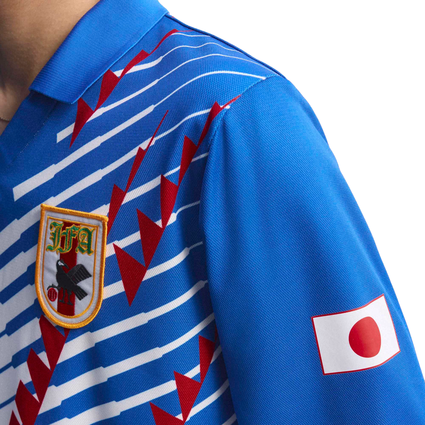 Close-up of a blue Japan national football jersey with white horizontal stripes, red jagged graphics, the JFA crest on the chest, and the Japanese flag on the sleeve.