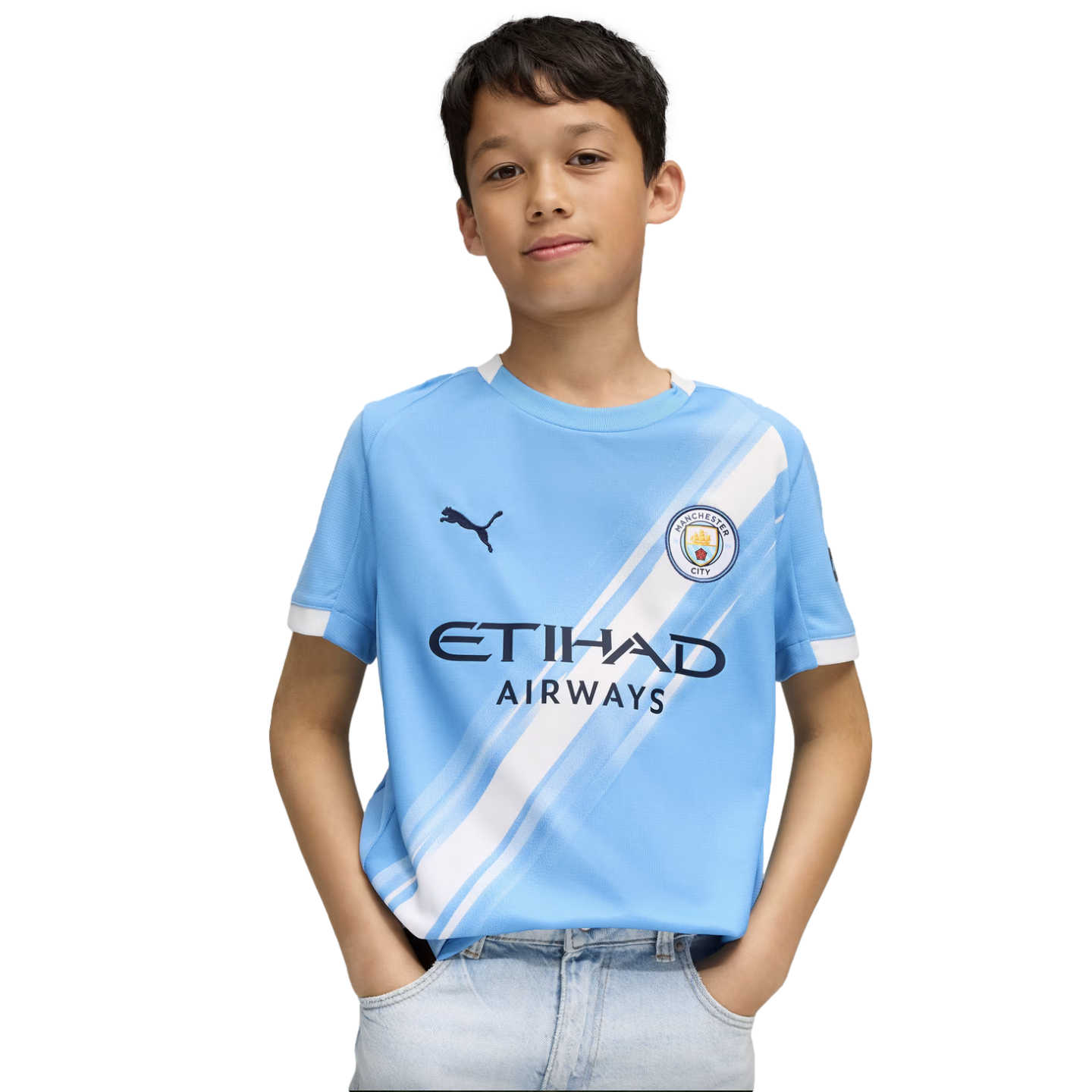 A young boy wearing a light blue Manchester City soccer jersey and light-wash jeans, standing with his hands in his pockets against a white background.