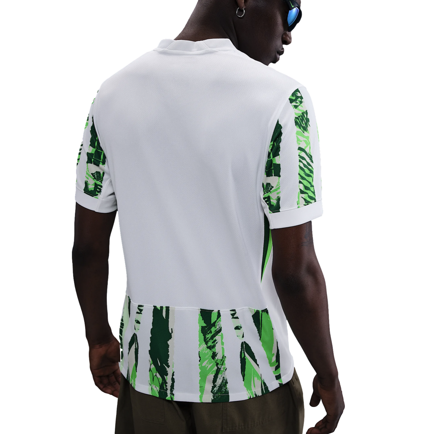 Back view of a person in a white short-sleeved athletic jersey with green abstract patterns on the sleeves and hem, worn with dark trousers against a white background.
