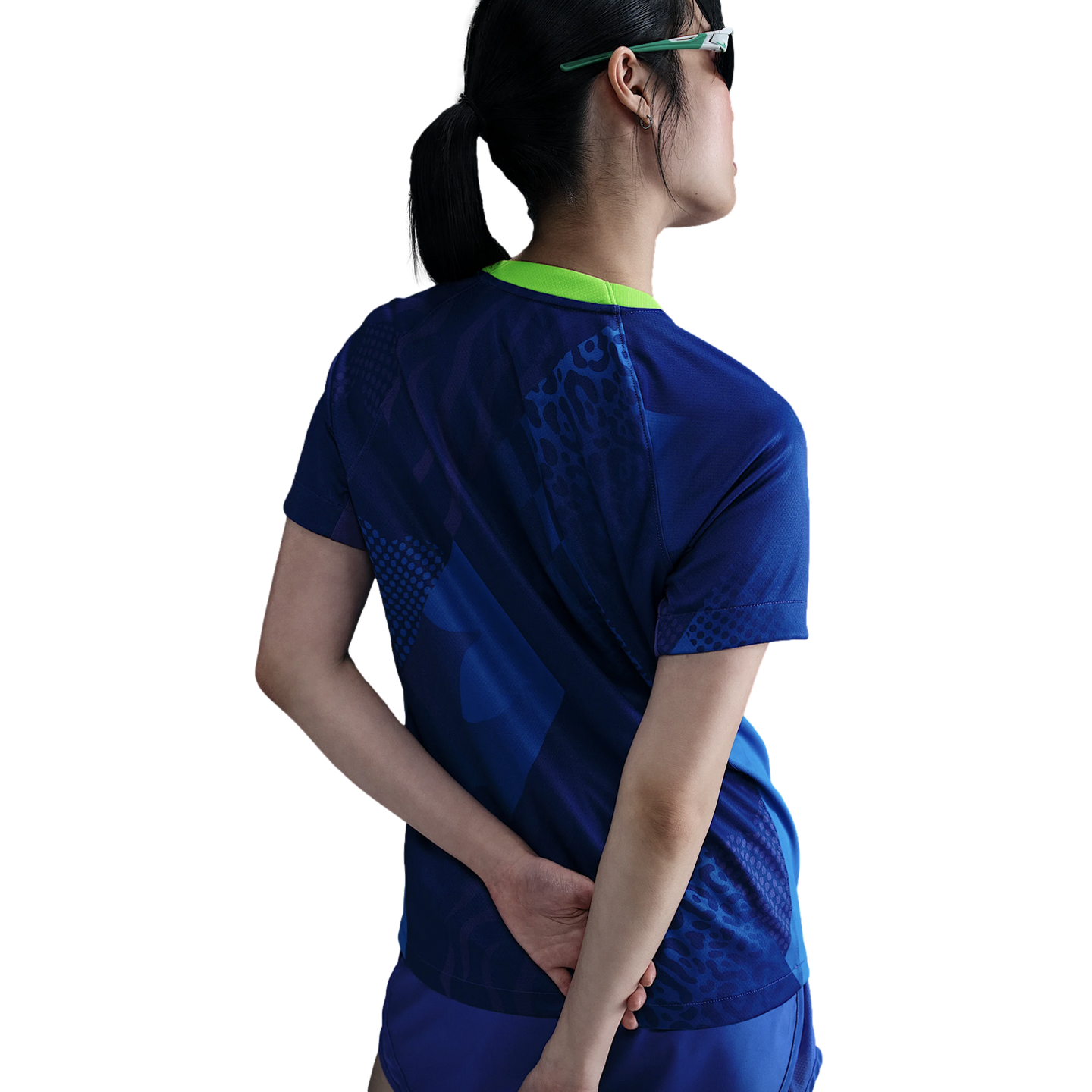 Rear view of a person wearing a blue patterned athletic shirt with a lime green collar and matching sunglasses, hands behind their back against a white background.
