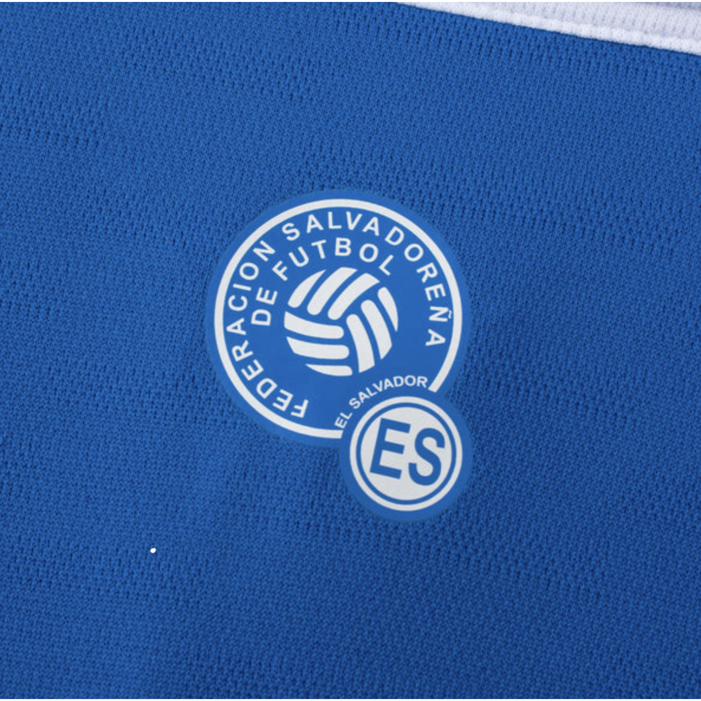 Close-up of the Federacion Salvadoreña de Futbol logo on a blue sports jersey. The white circular emblem features a soccer ball and the initials "ES".