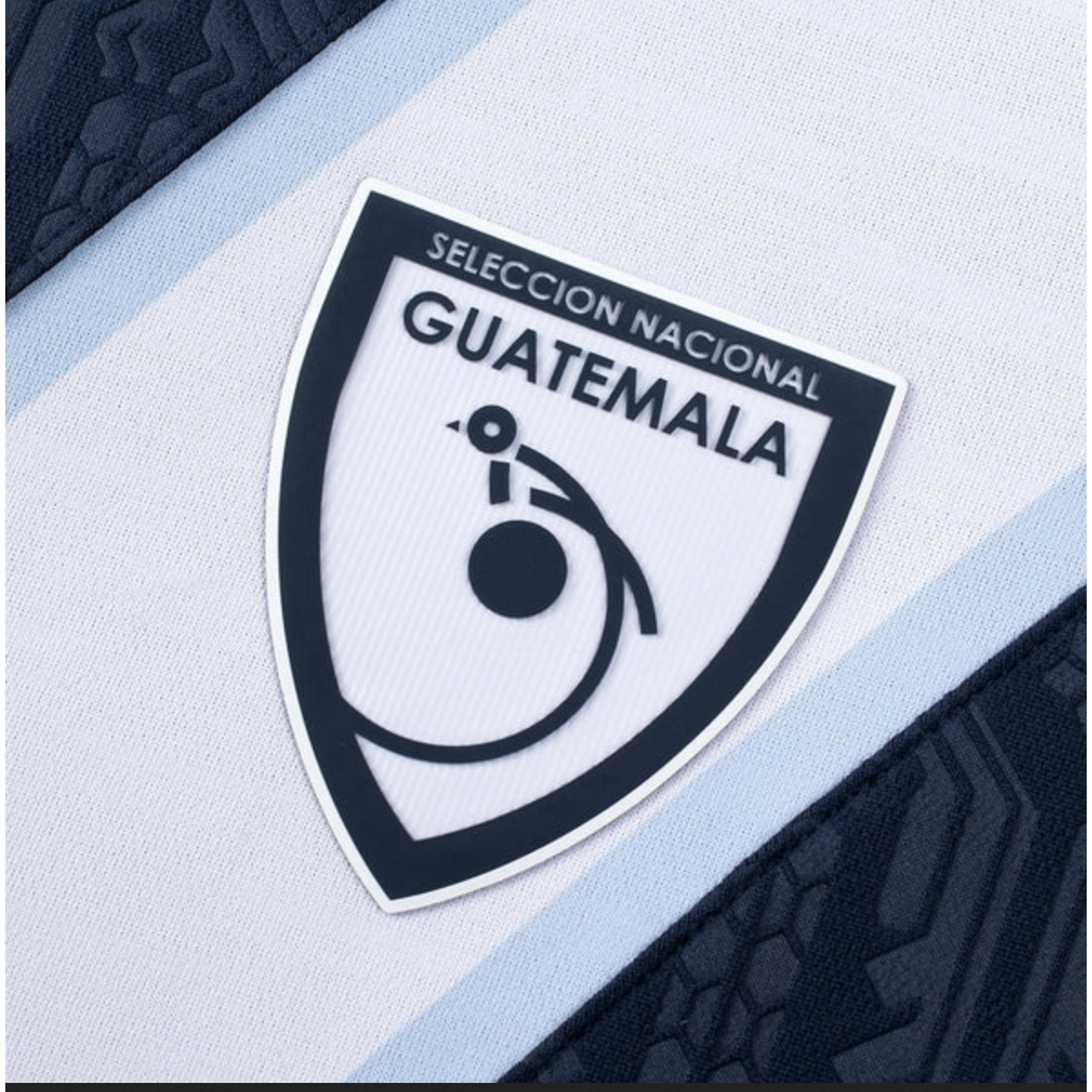 Close-up of the Guatemala national team crest on a white and blue jersey, featuring a stylized quetzal logo and the words "SELECCIÓN NACIONAL GUATEMALA."
