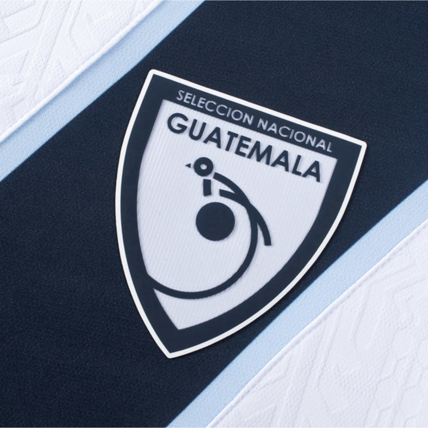Close-up of the Guatemala national soccer team shield crest on a navy and light blue jersey, featuring a stylized quetzal bird.