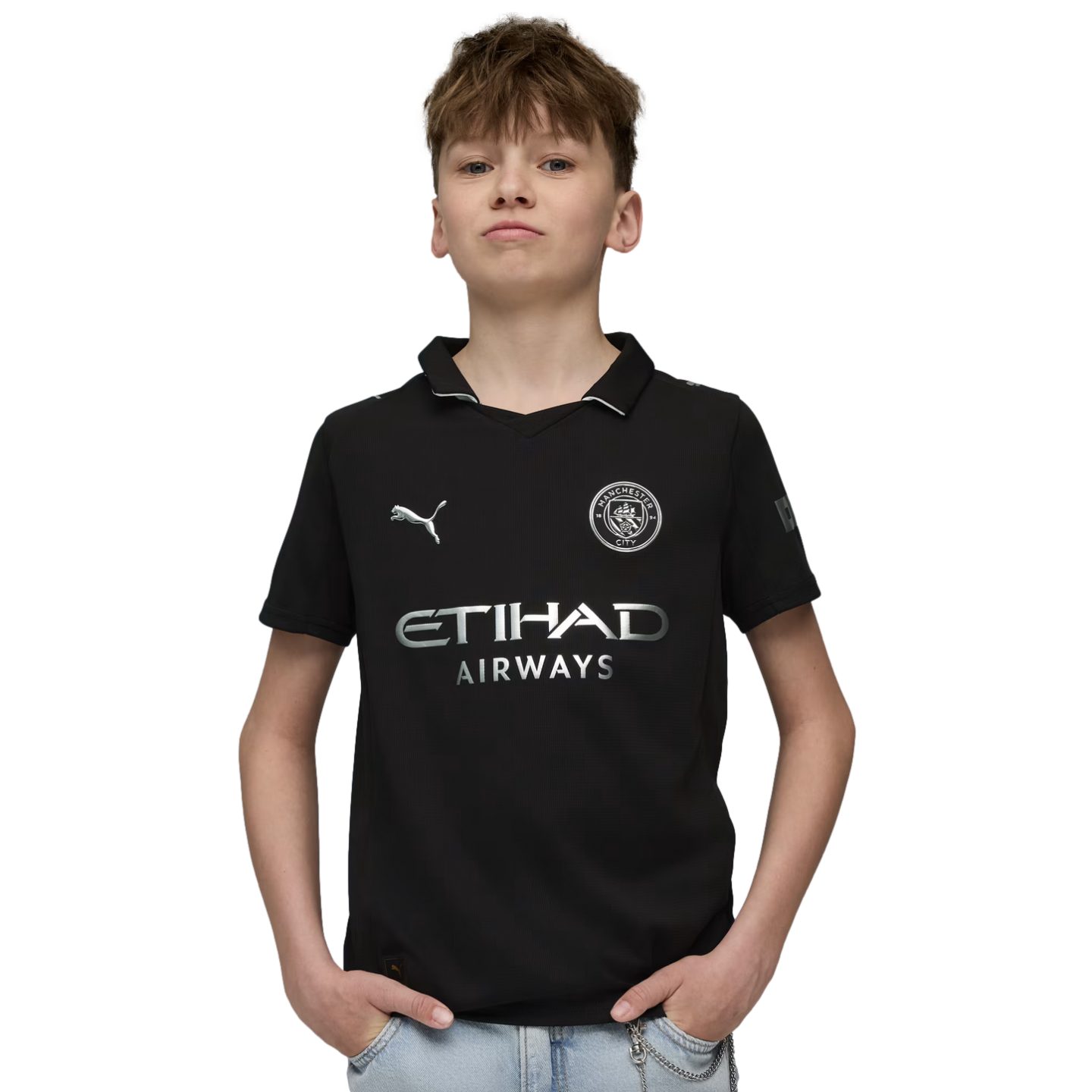 A young boy with light brown hair wears a black Manchester City soccer jersey and light blue jeans, posing with his hands in his pockets against a solid black background.