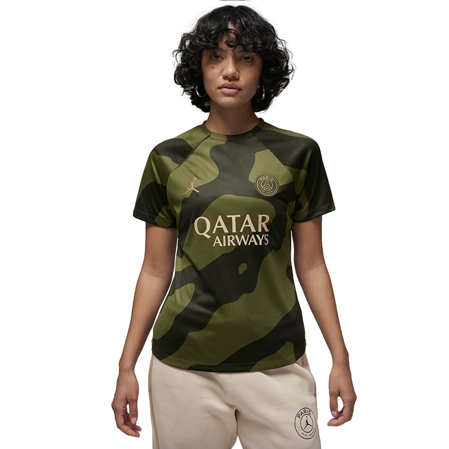 A woman with short curly hair models a green camouflage Jordan x PSG soccer jersey and cream joggers against a black background.