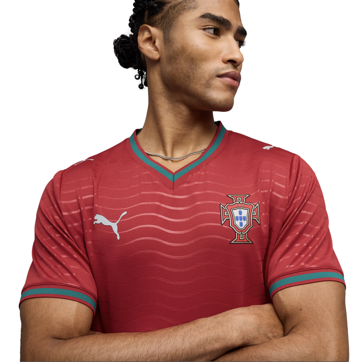 Man with braided hair in a red Portugal Puma soccer jersey, arms crossed, looking sideways against a black background.