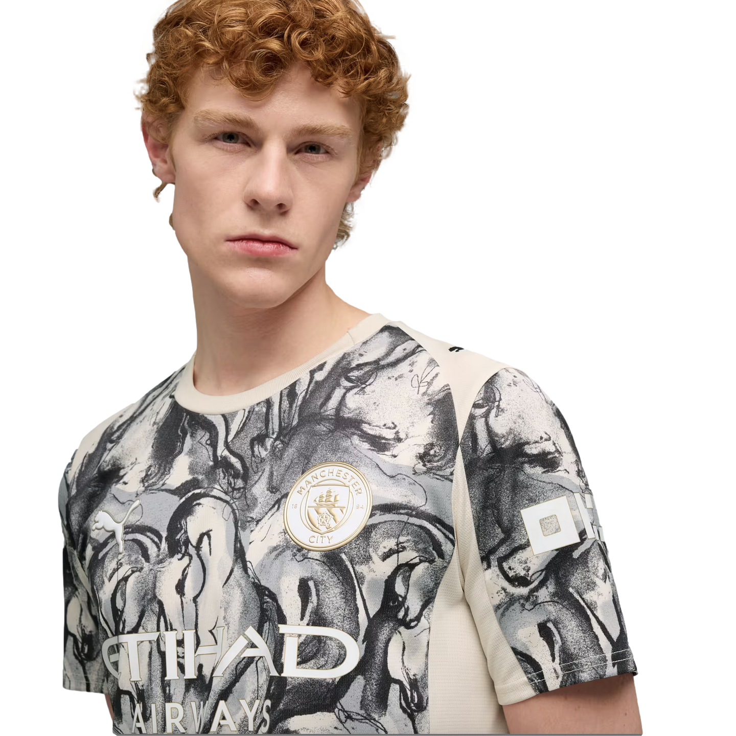A young man with curly red hair wearing a grey and white patterned Manchester City soccer jersey against a black background.