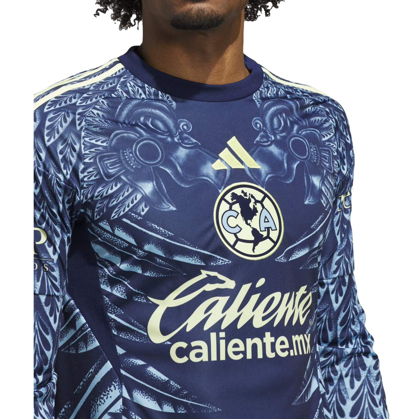 Close-up of a navy blue Club América soccer jersey with a light blue eagle pattern, yellow Adidas logo, and "Caliente" sponsor logo.