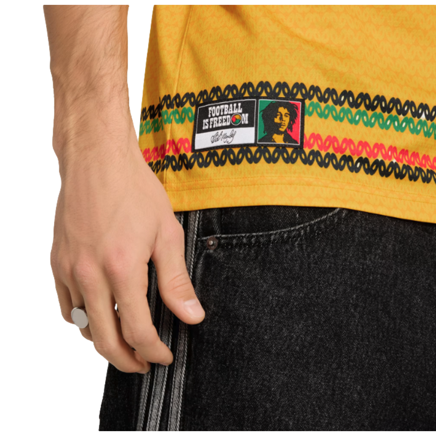 Close-up of a yellow jersey with a Bob Marley "Football is Freedom" patch and knit-patterned stripes, worn with black striped denim. A silver ring is visible on the person's hand.