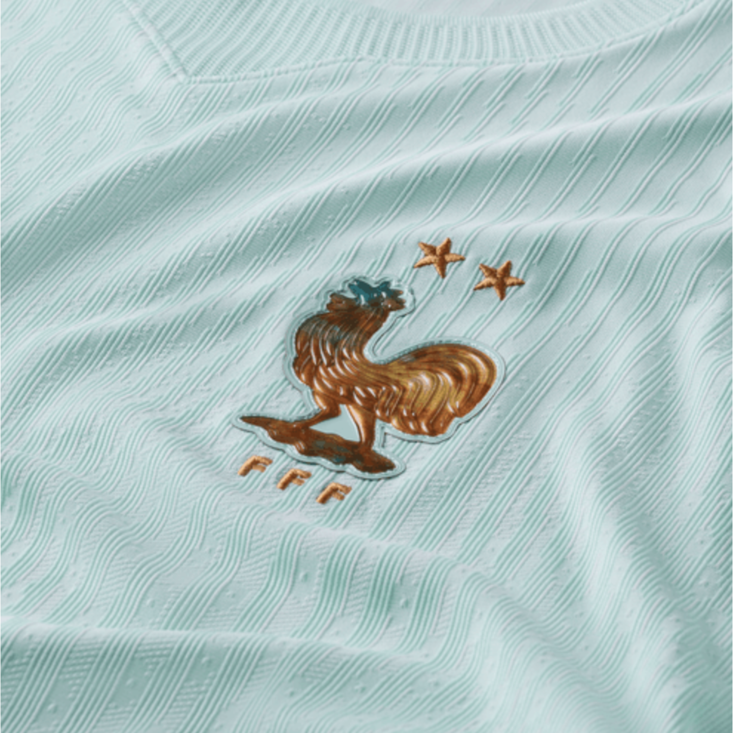 Close-up of a light blue textured football jersey featuring the golden rooster crest of the French Football Federation (FFF) and two stars.