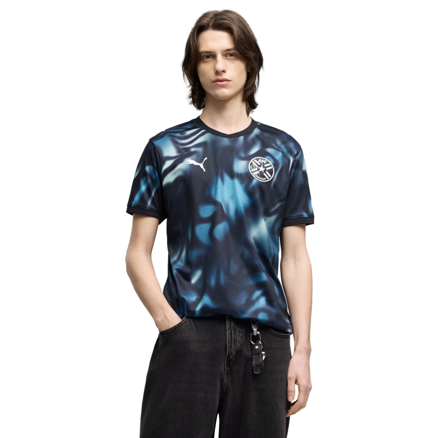 A man with brown hair wears a blue and black abstract Puma jersey with the Paraguay football crest and black jeans against a solid black background.