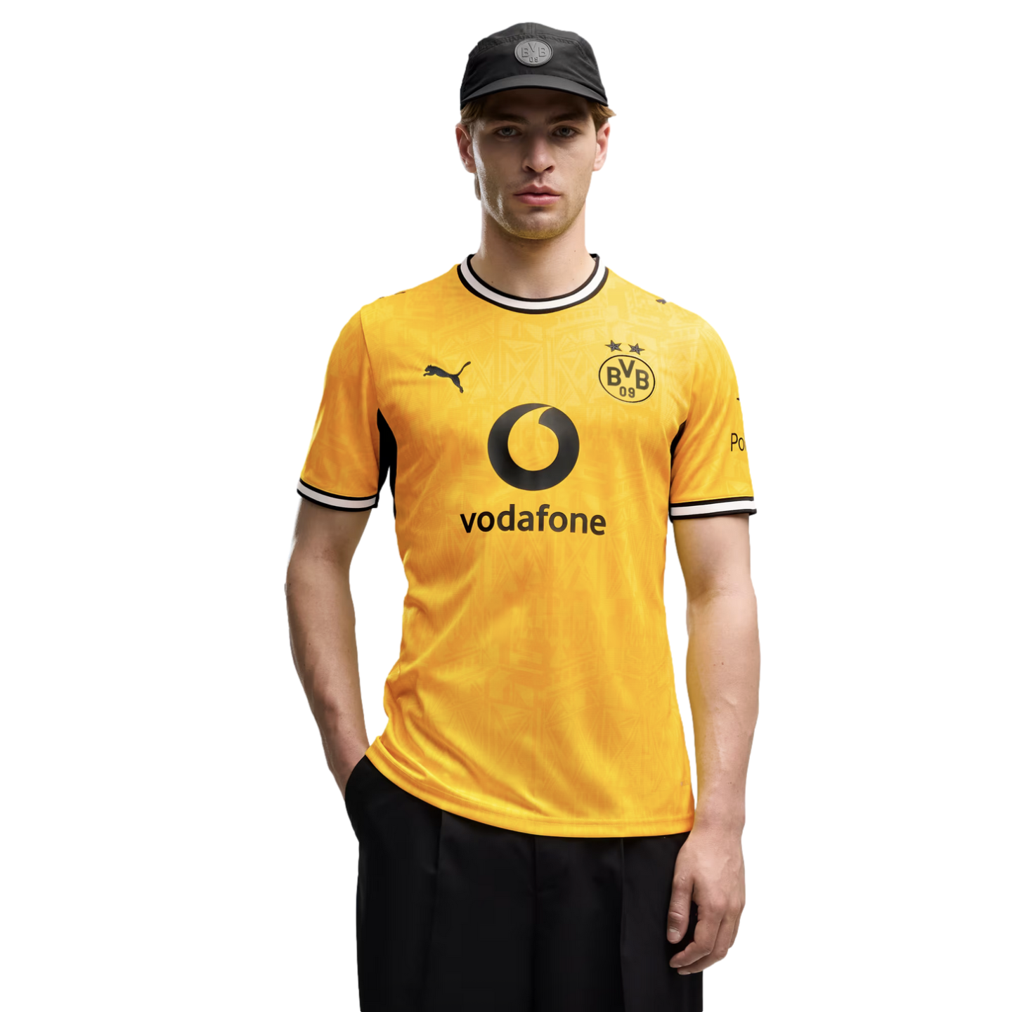 A man in a yellow Borussia Dortmund soccer jersey with Vodafone and Puma logos, wearing a black cap and posed against a solid black background.
