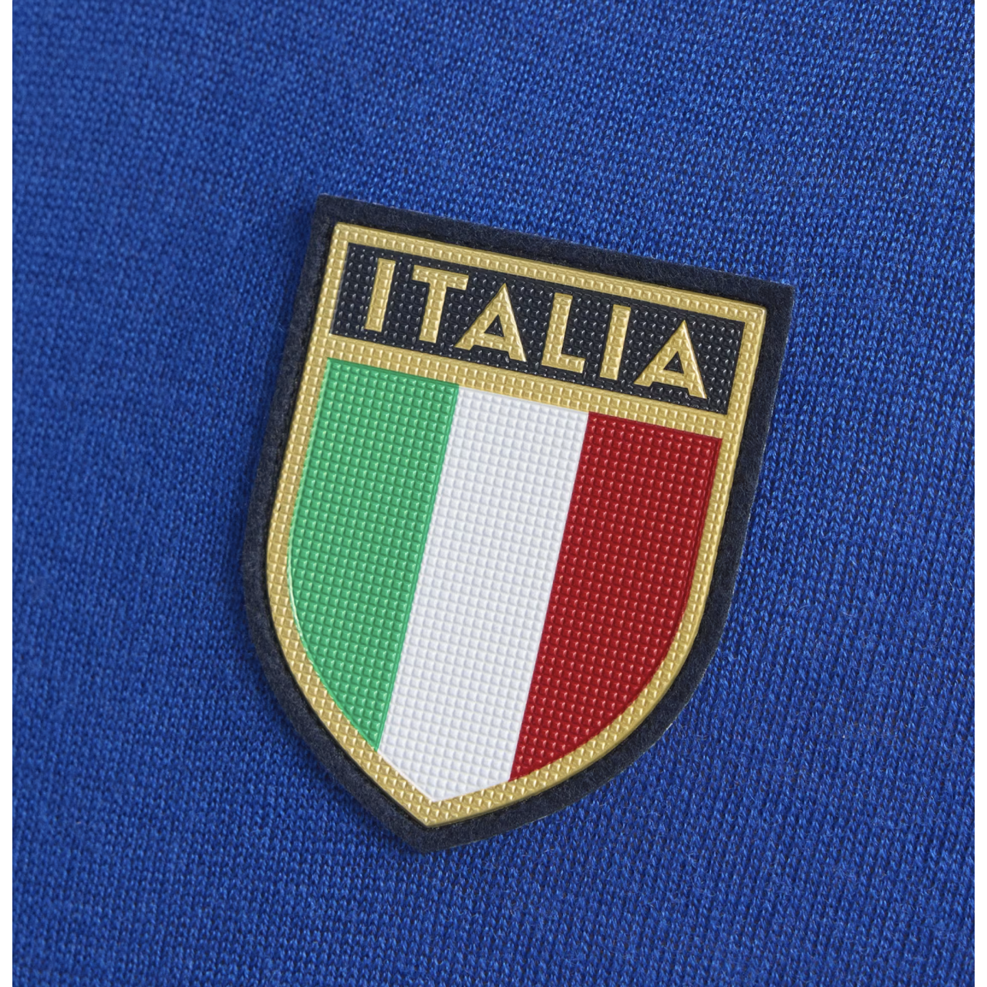 A close-up of a shield-shaped "ITALIA" patch on blue fabric, featuring gold text above vertical green, white, and red stripes.
