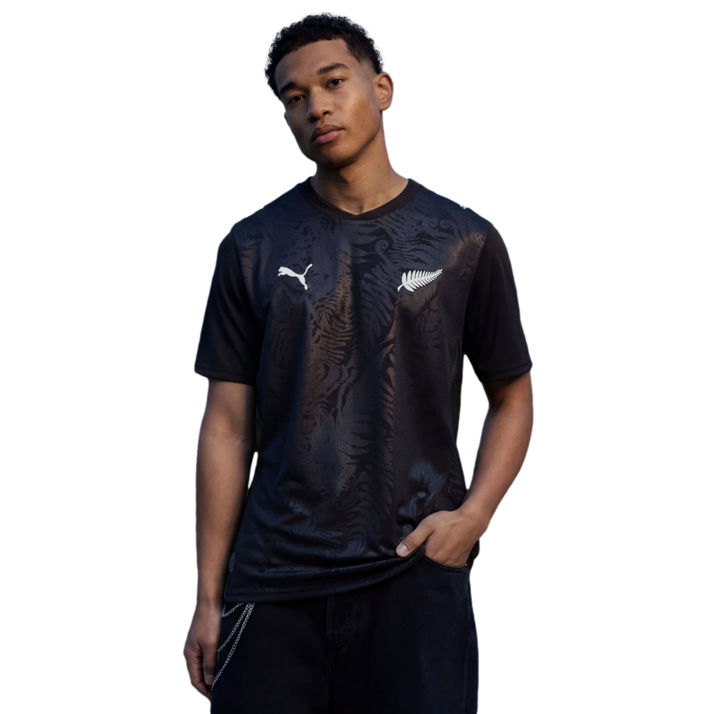 A young man poses against a black background, wearing a black Puma t-shirt featuring a tonal fern pattern and the white New Zealand Silver Fern logo.